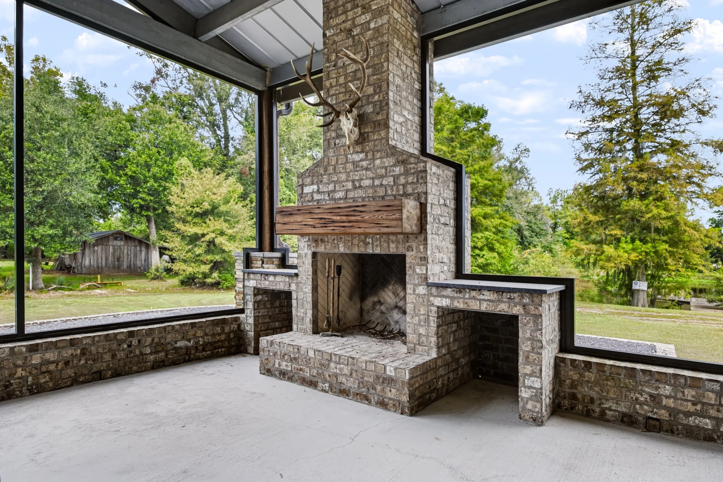 Interior view of a stone fireplace with a mounted deer skull and antlers, surrounded by large windows showing green trees and blue sky outside.