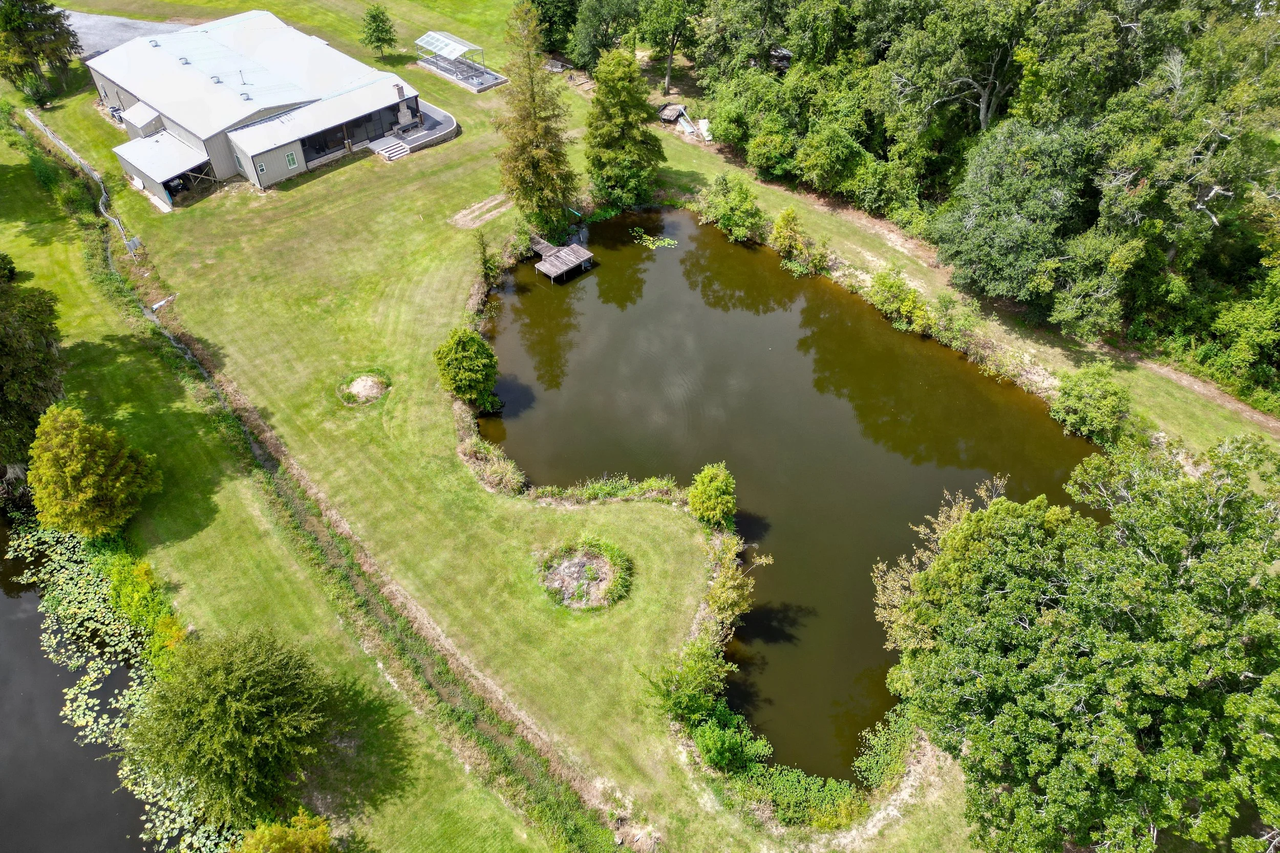 An aerial view of a house with a metal roof located beside a pond, surrounded by green grass, trees, and a wooded area.