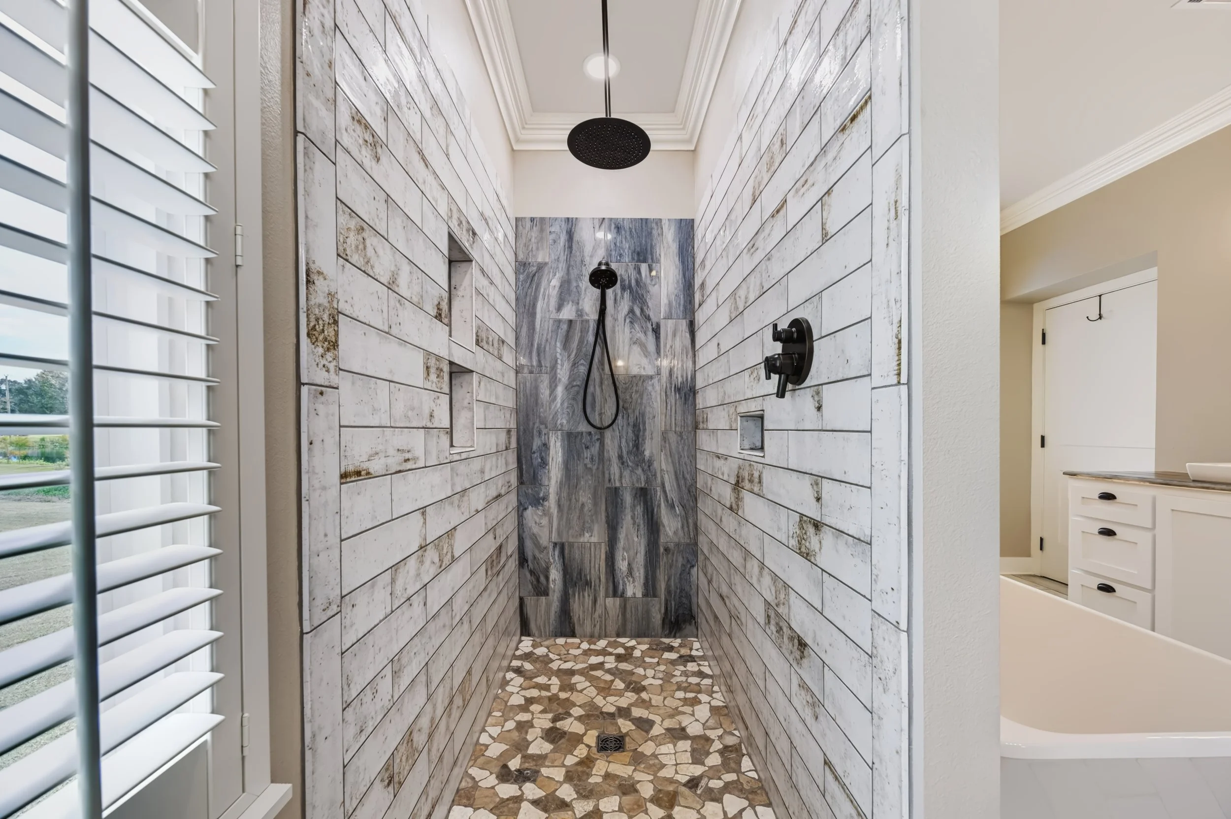 Indoor shower with gray and white marble tiles on the wall, pebble floor, and black rainfall showerheads.