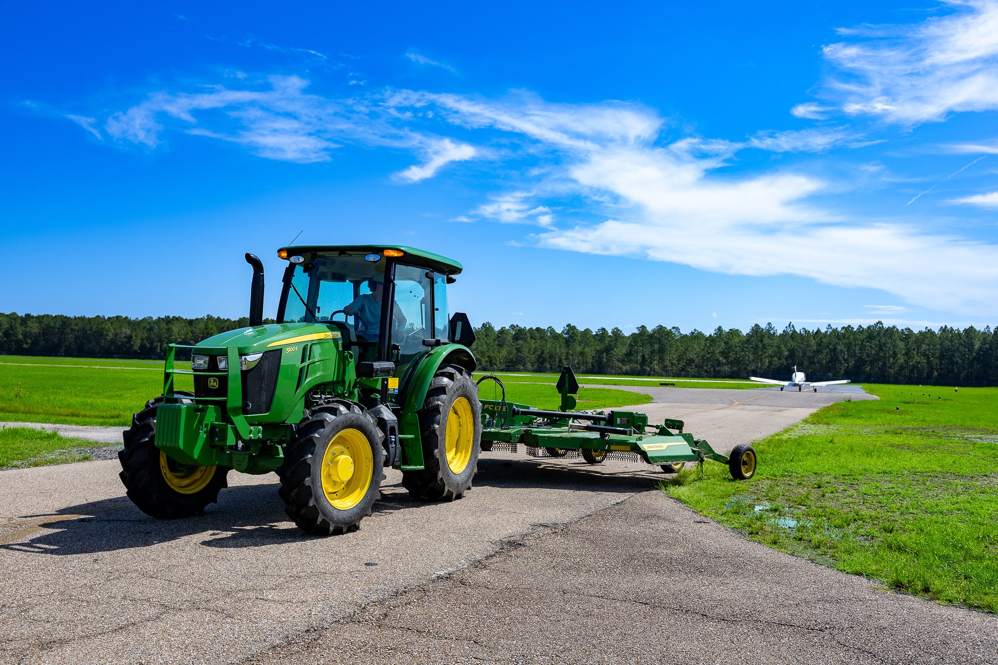 A green John Deere tractor with yellow wheels and a black exhaust pipe in a grassy area, with a clear blue sky and some clouds above, and an airplane parked in the background.