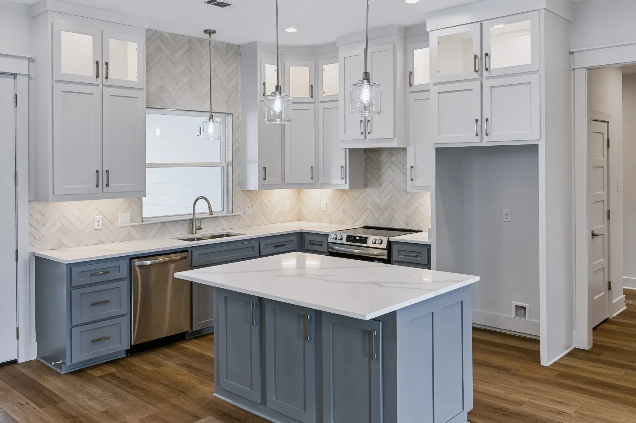 Modern kitchen with white upper cabinets, gray lower cabinets, a marble countertop island, and a herringbone-patterned tile backsplash. Two pendant lights hang above the island, and there is a window above the sink.