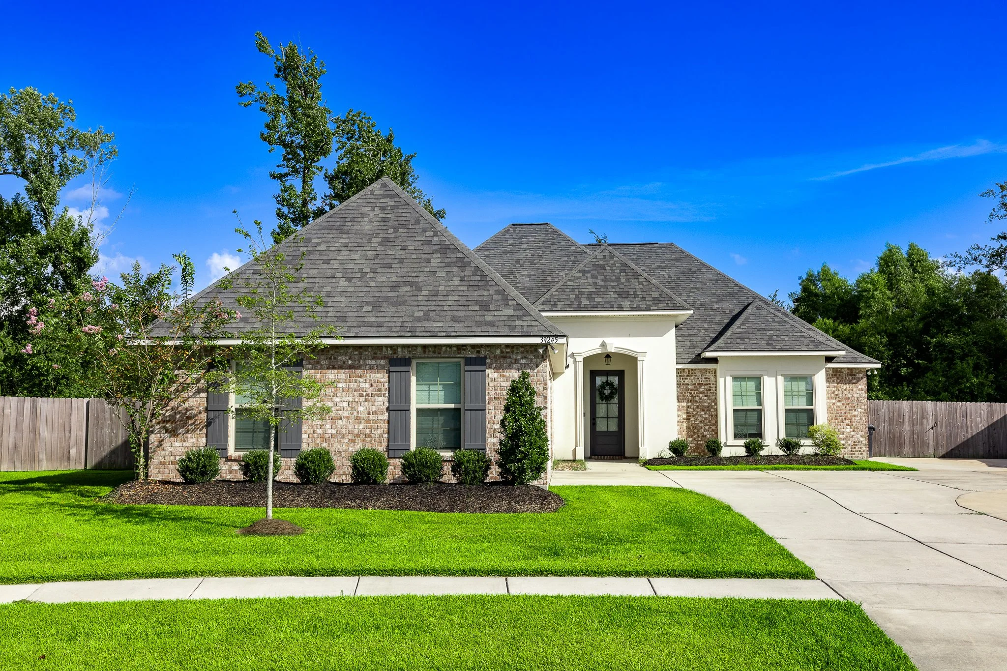 Front view of a house with brick and white siding, a gray shingle roof, a black front door, and a green lawn with small bushes and a tree, under a clear blue sky.