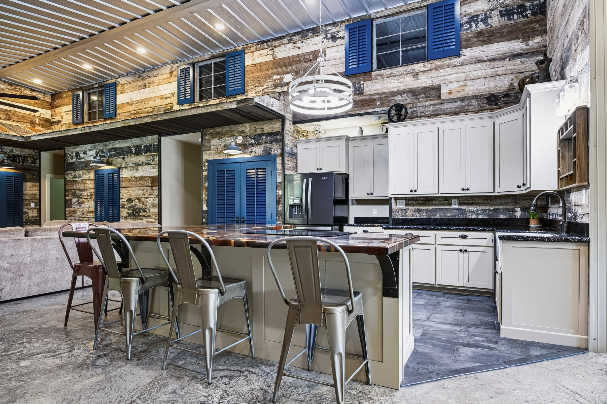 Interior of a rustic kitchen with wood-paneled walls, white cabinets, a black refrigerator, and a large wooden dining table with metal chairs.