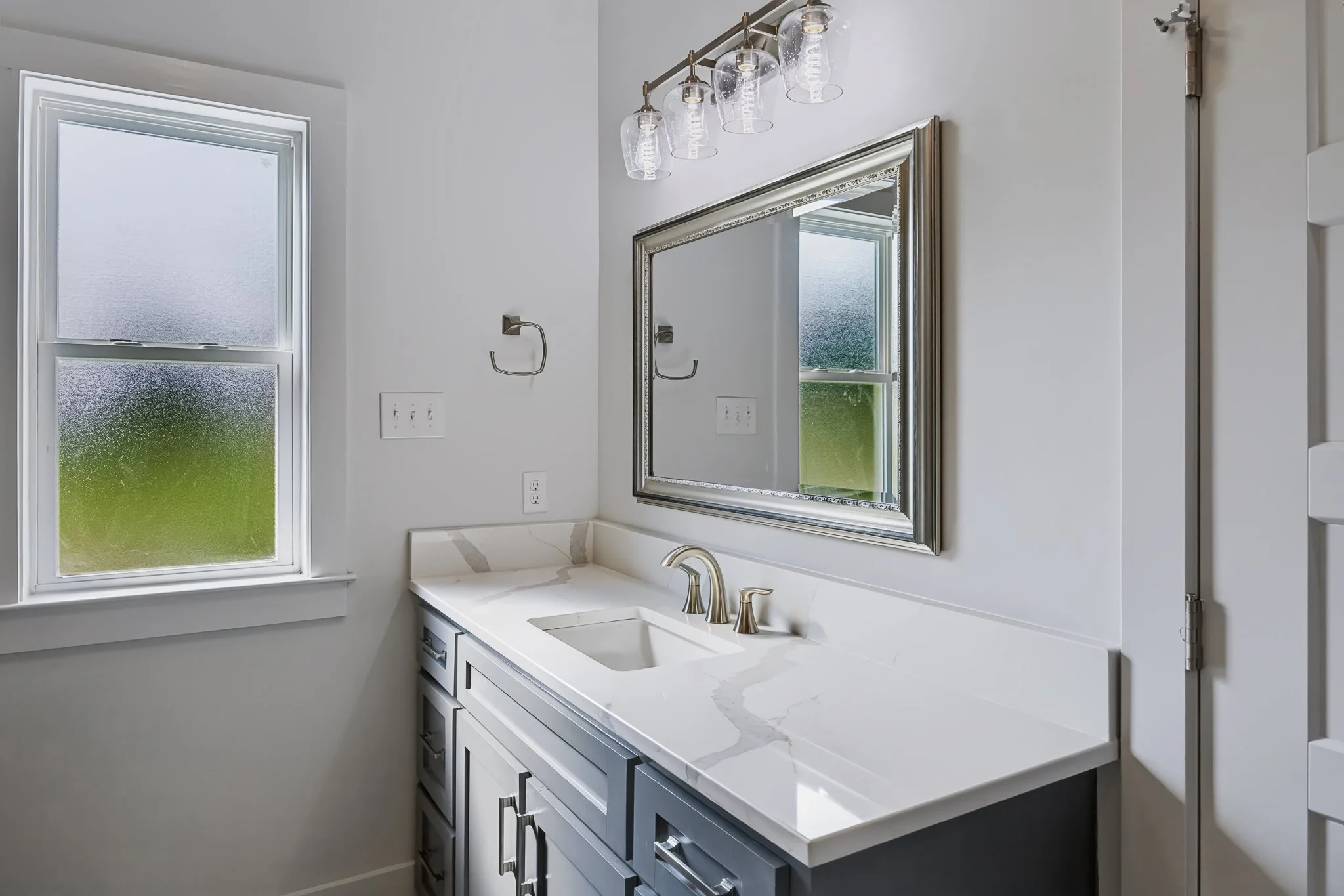 Bathroom with white cabinets, marble countertop, stainless steel faucet, mirror, frosted window, wall-mounted light fixture with glass shades, and a towel hook.