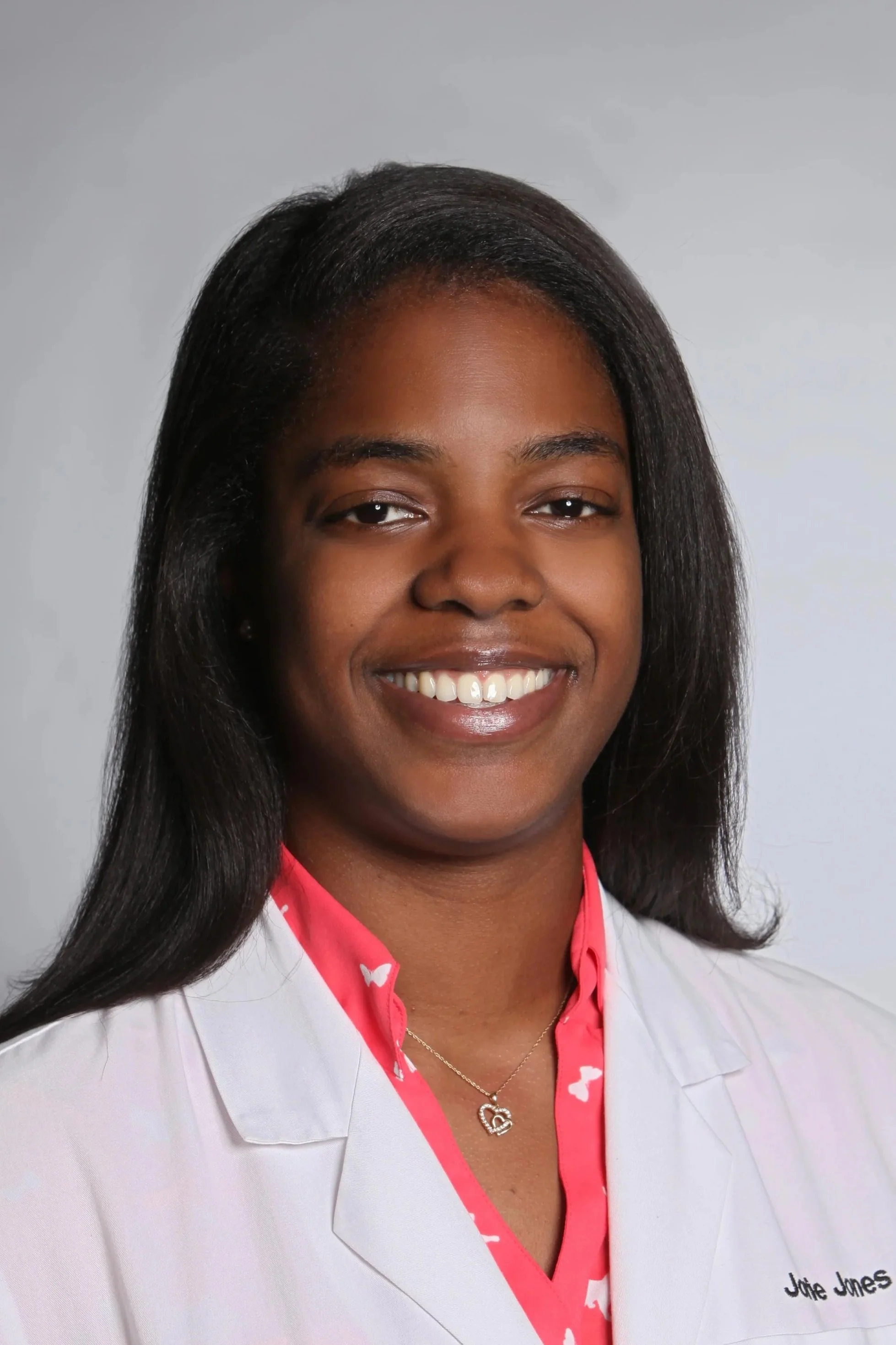 Close-up of a smiling woman with dark hair, wearing a white coat over a pink patterned shirt, against a plain gray background.