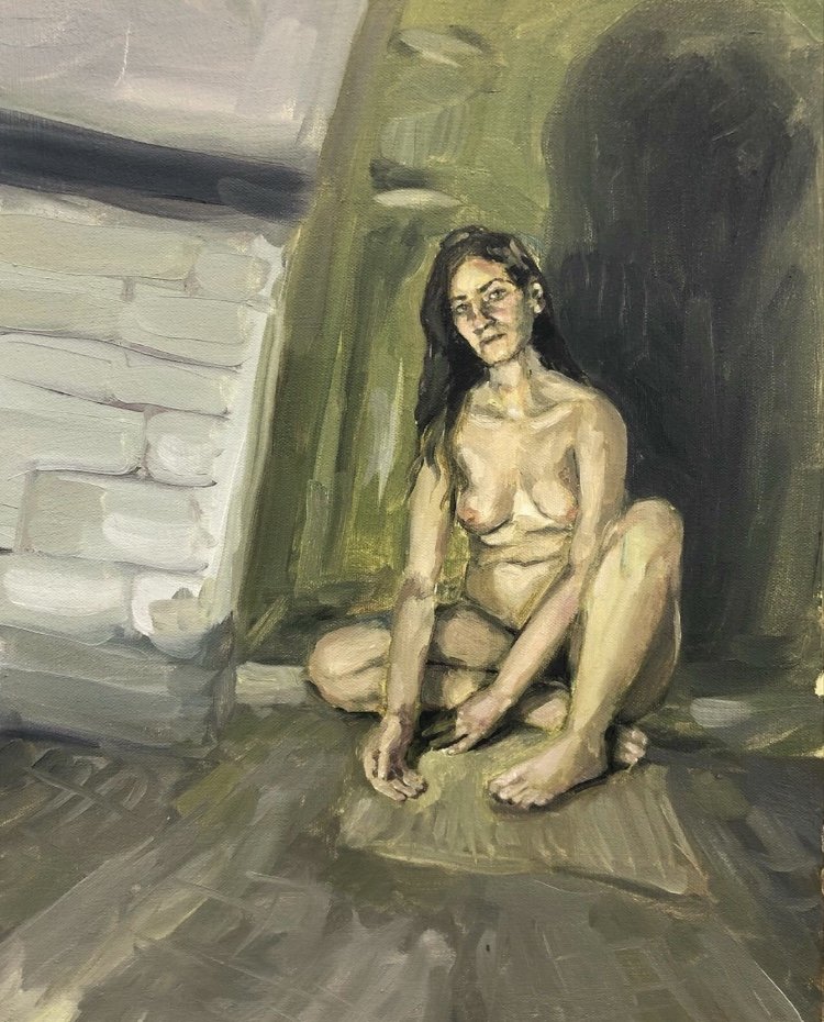 A nude woman with long dark hair sitting on the floor of a rustic room with a brick wall and wooden paneling, illuminated by a light source from above, casting a dark shadow behind her.