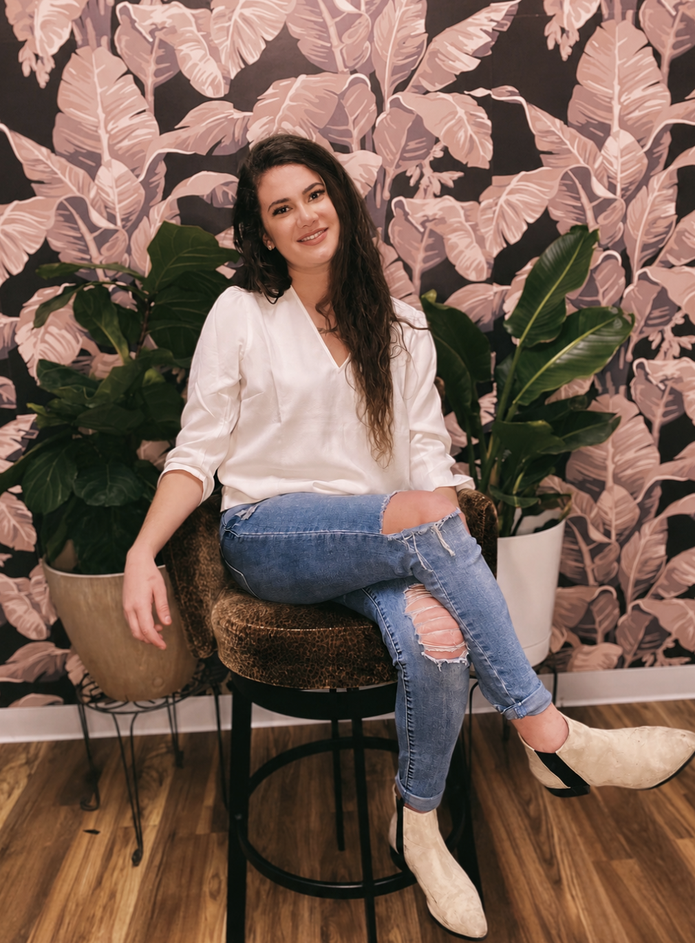 A woman with long dark hair sitting on a patterned brown chair against a pink and dark background with tropical leaves, flanked by two large green potted plants.