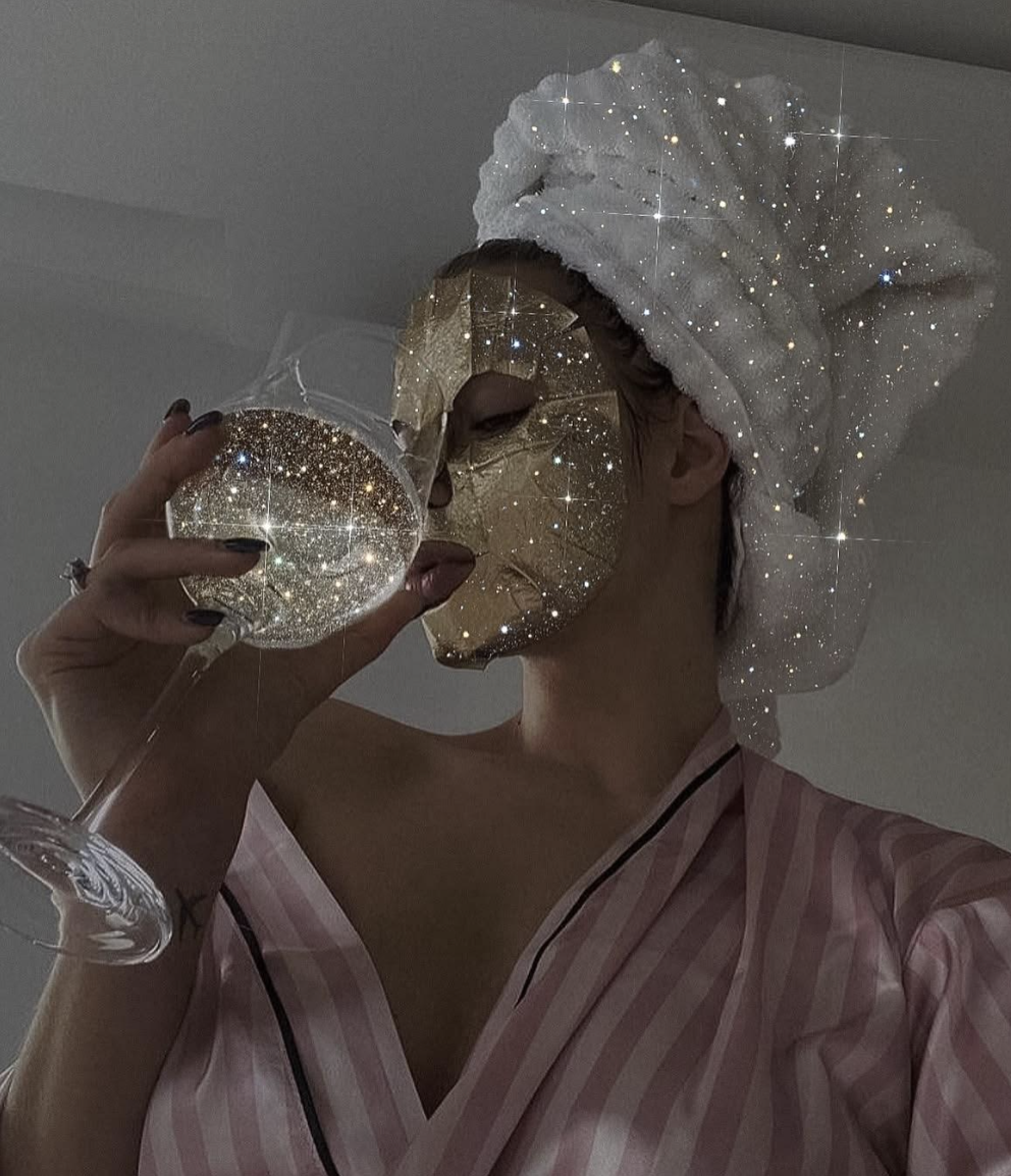 Person wearing a white towel turban, sipping wine from a glass, with a gold mask covering their face, in a dressing gown, and a sparkle overlay effect.