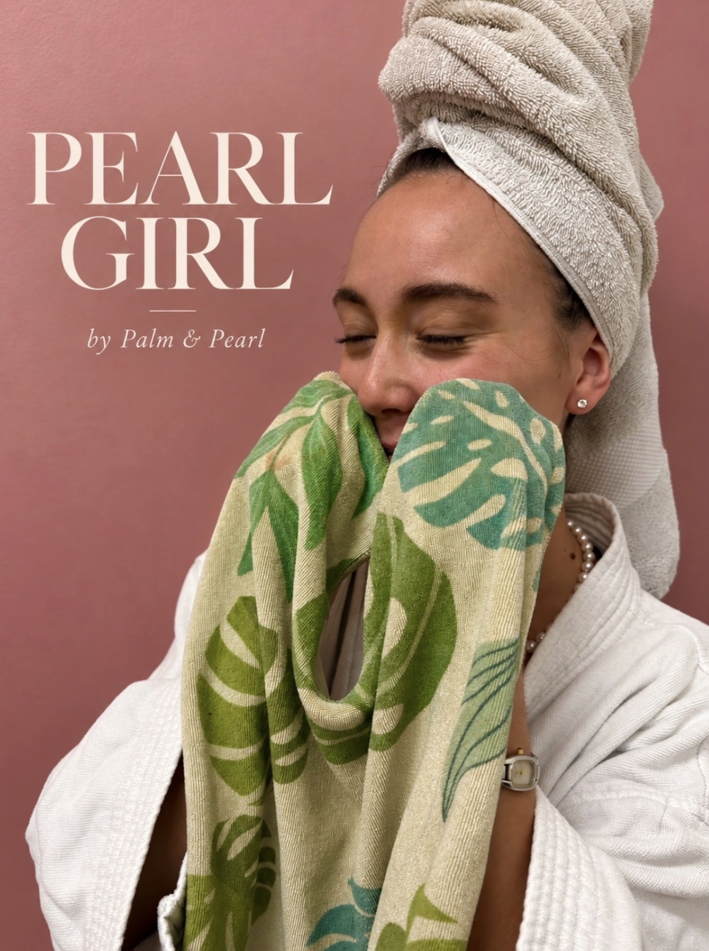 A woman with a towel wrapped around her head, smiling with eyes closed, holding a green and beige patterned towel close to her face, against a pink background.