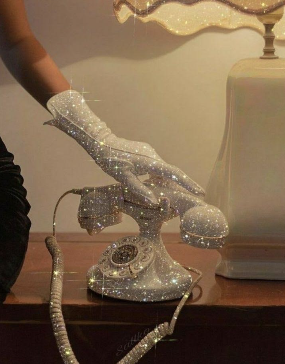 A vintage rotary telephone decorated with sparkling, glittering embellishments sitting on a wooden surface next to a lamp with a beige base and a shade with a swirling design.