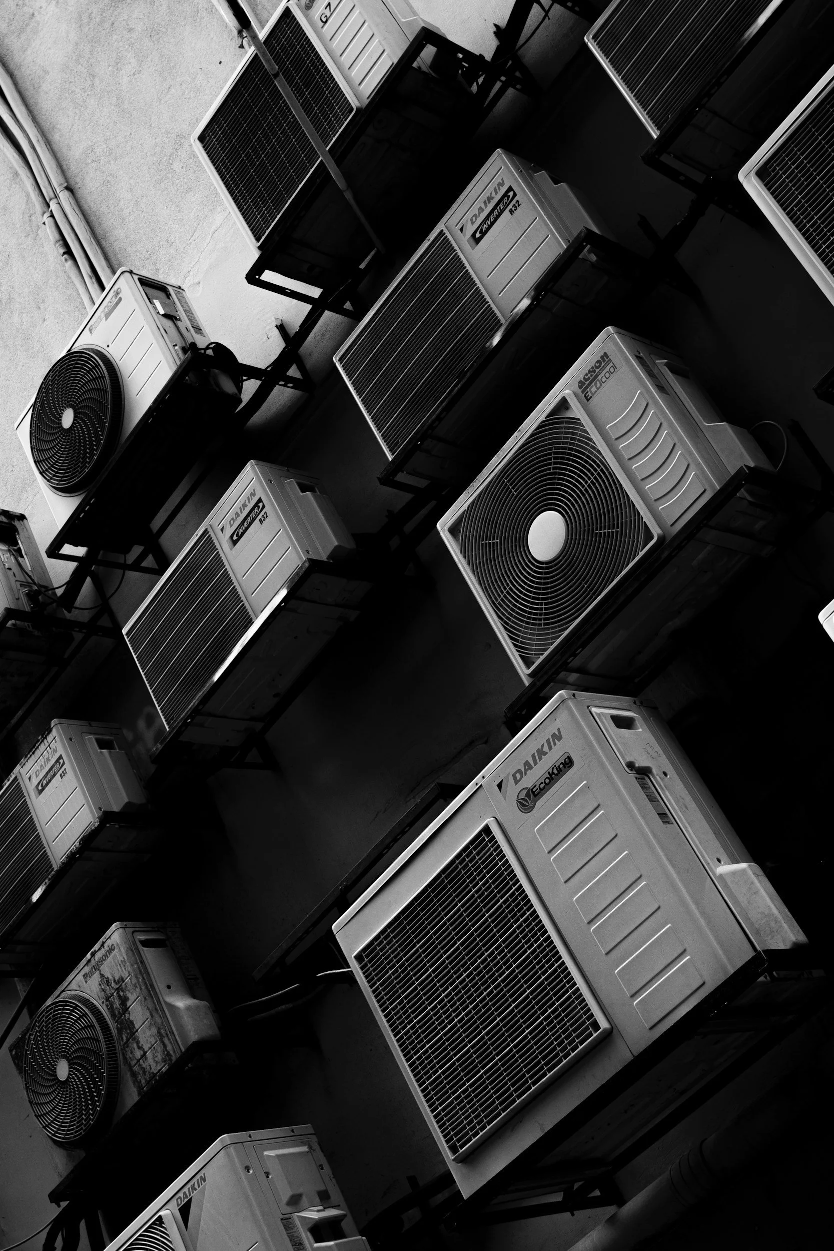 Black and white photo of multiple outdoor air conditioning units mounted on a wall, arranged in rows and supported by metal frames.