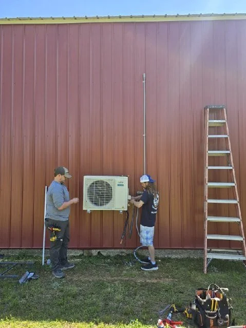 Two workers installing or servicing an outdoor air conditioning unit on the exterior wall of a red building, with a ladder leaning against the wall and tools on the ground.