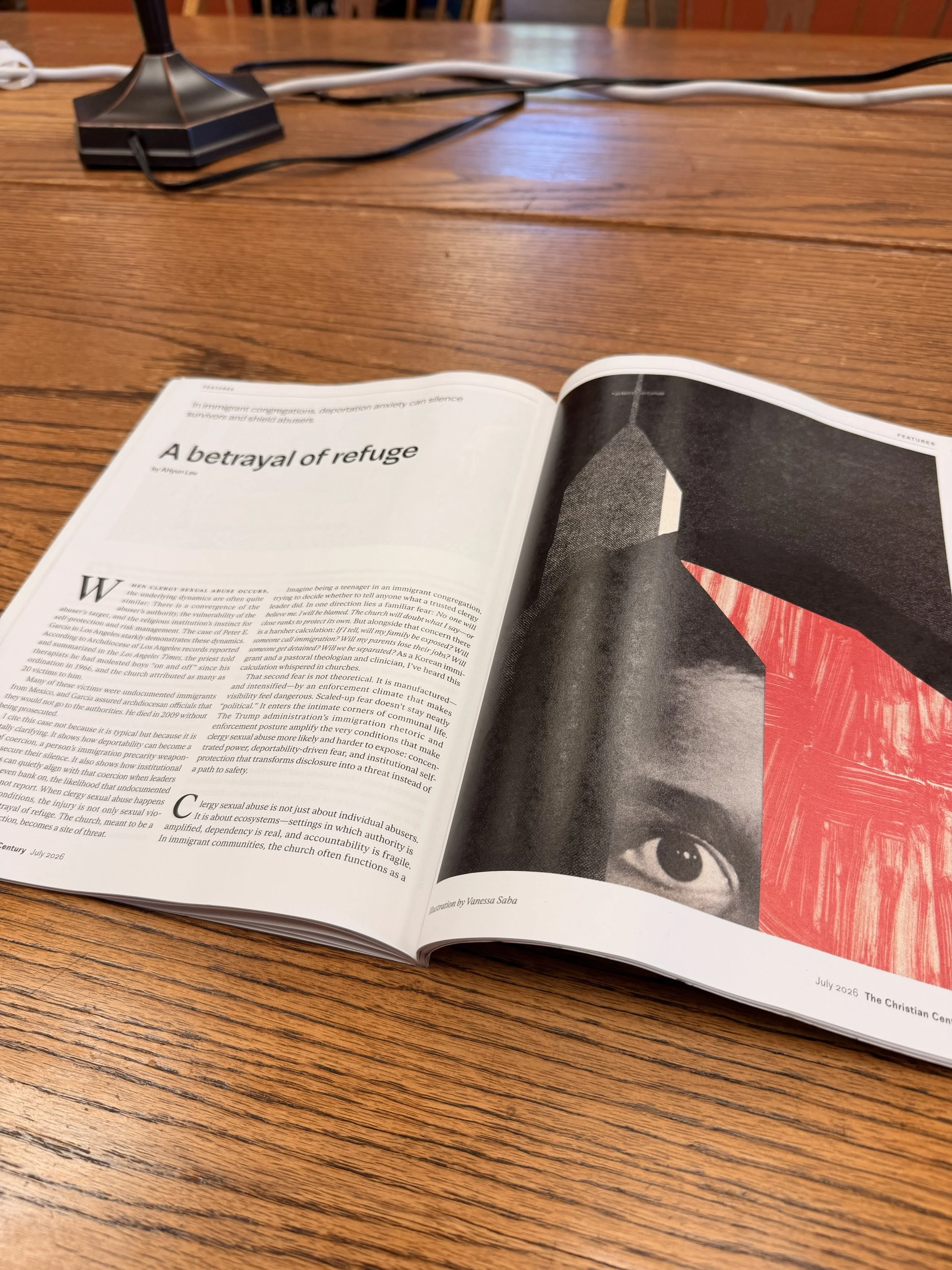 An open magazine on a wooden table showing an article titled "A betrayal of refuge" with a black-and-white image of a church with a cross on top, featuring a close-up of a person's eye at the bottom of the page.
