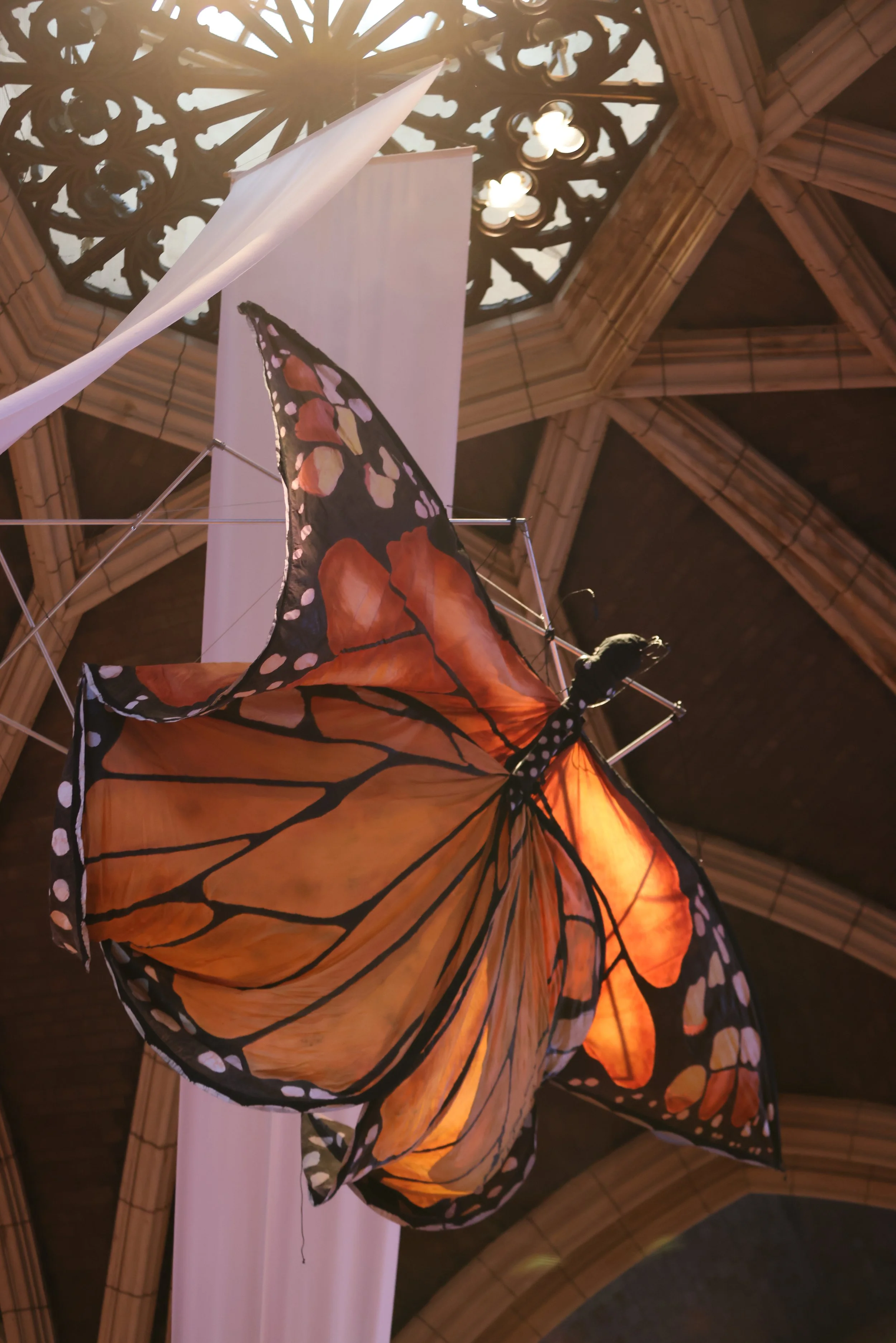 A butterfly-shaped kite with orange, black, and white patterns hanging from a display stand inside a building with wooden architecture and a stained glass window on the ceiling.