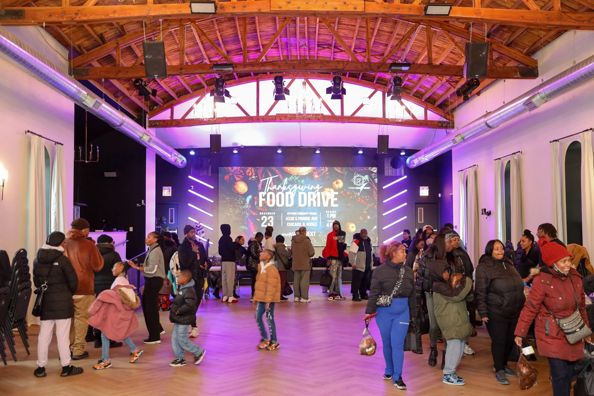 Crowd of people attending a Thanksgiving food drive event inside a large hall with a high wooden ceiling, purple lighting, and a large digital screen displaying event details.