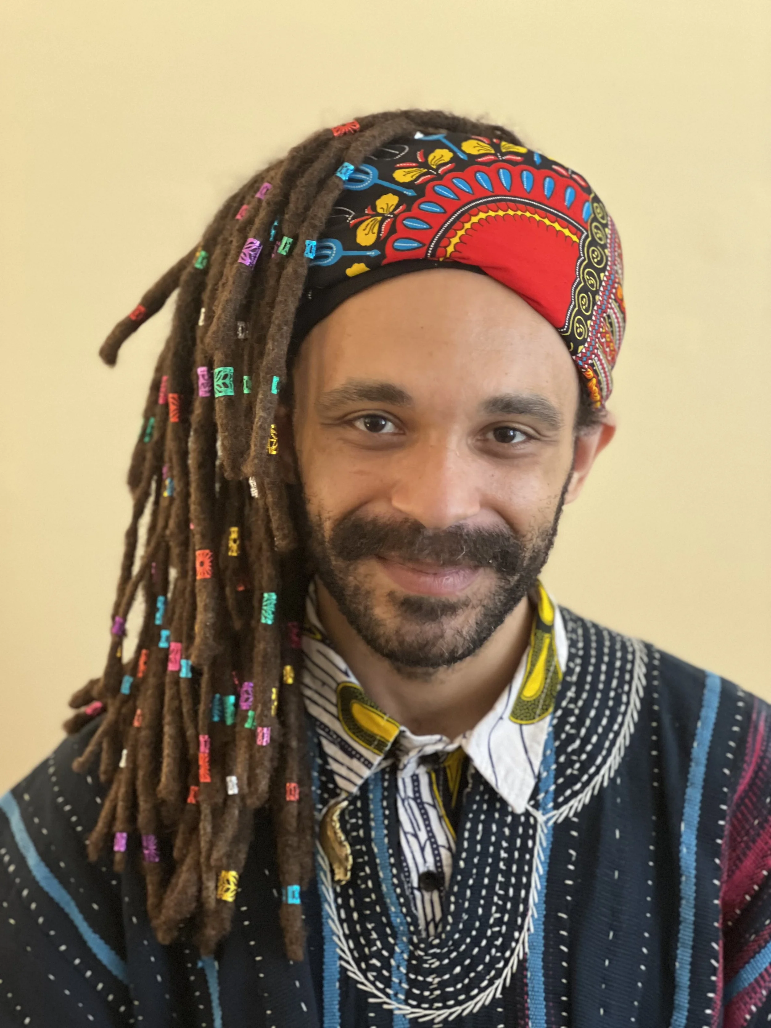 A person with dark skin and dreadlocks wearing a vibrant, patterned headband and a black sweater with colorful lines, smiling at the camera.