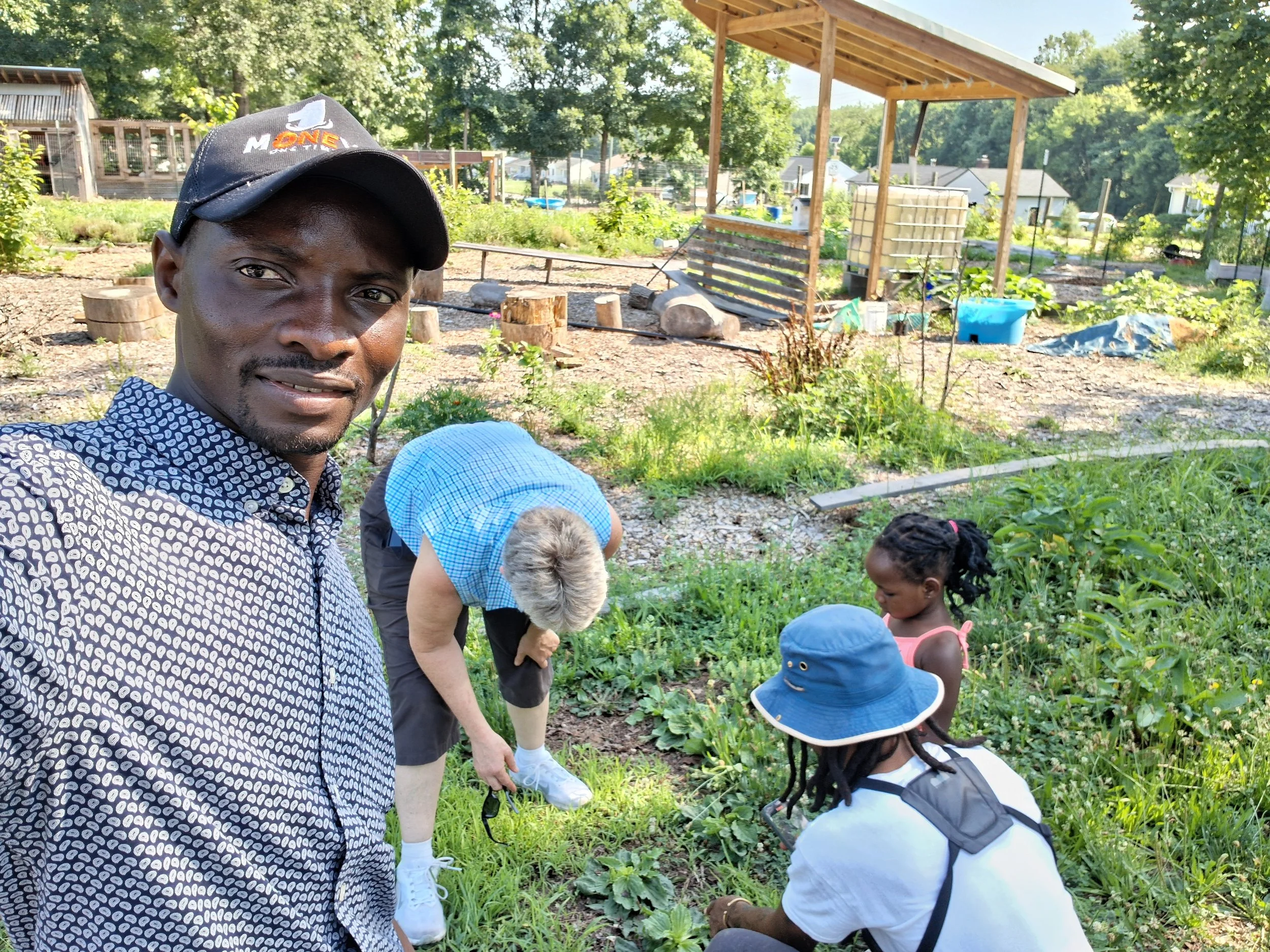 A man wearing a black cap with an orange logo and a patterned shirt is taking a selfie in a garden with children and adults tending to plants. The garden has trees, plants, and gardening supplies, with buildings and a fence in the background.