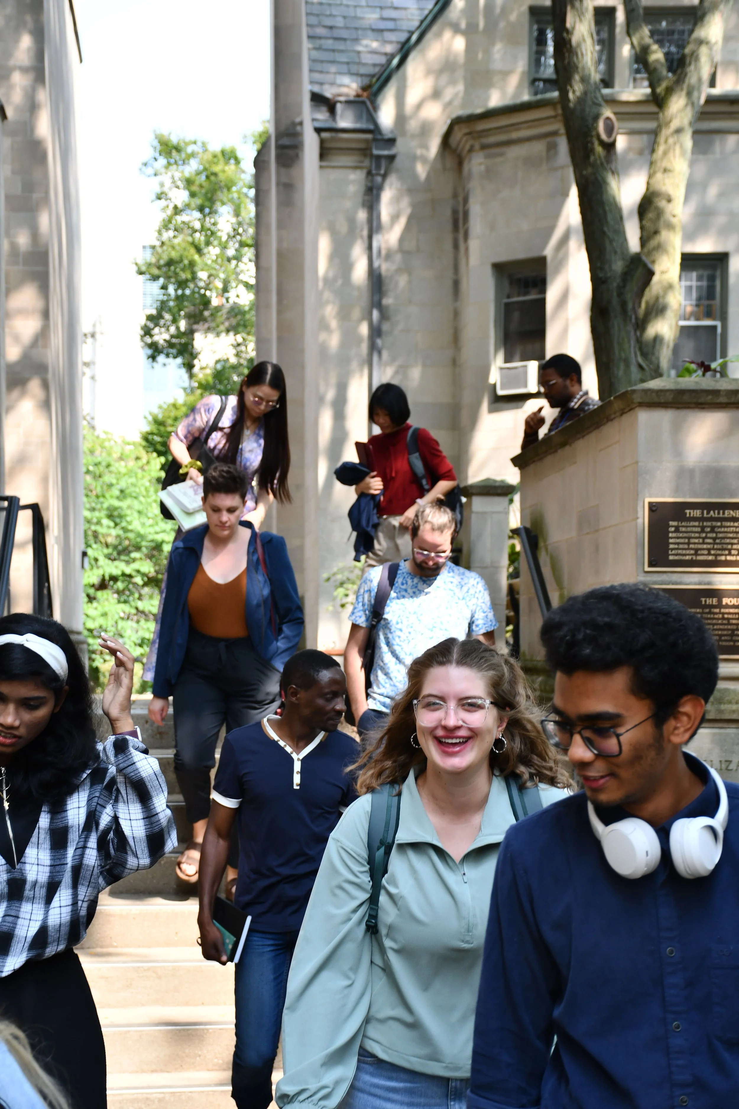 Group of diverse young adults walking down outdoor stone steps, smiling, with a stone building and trees in the background.