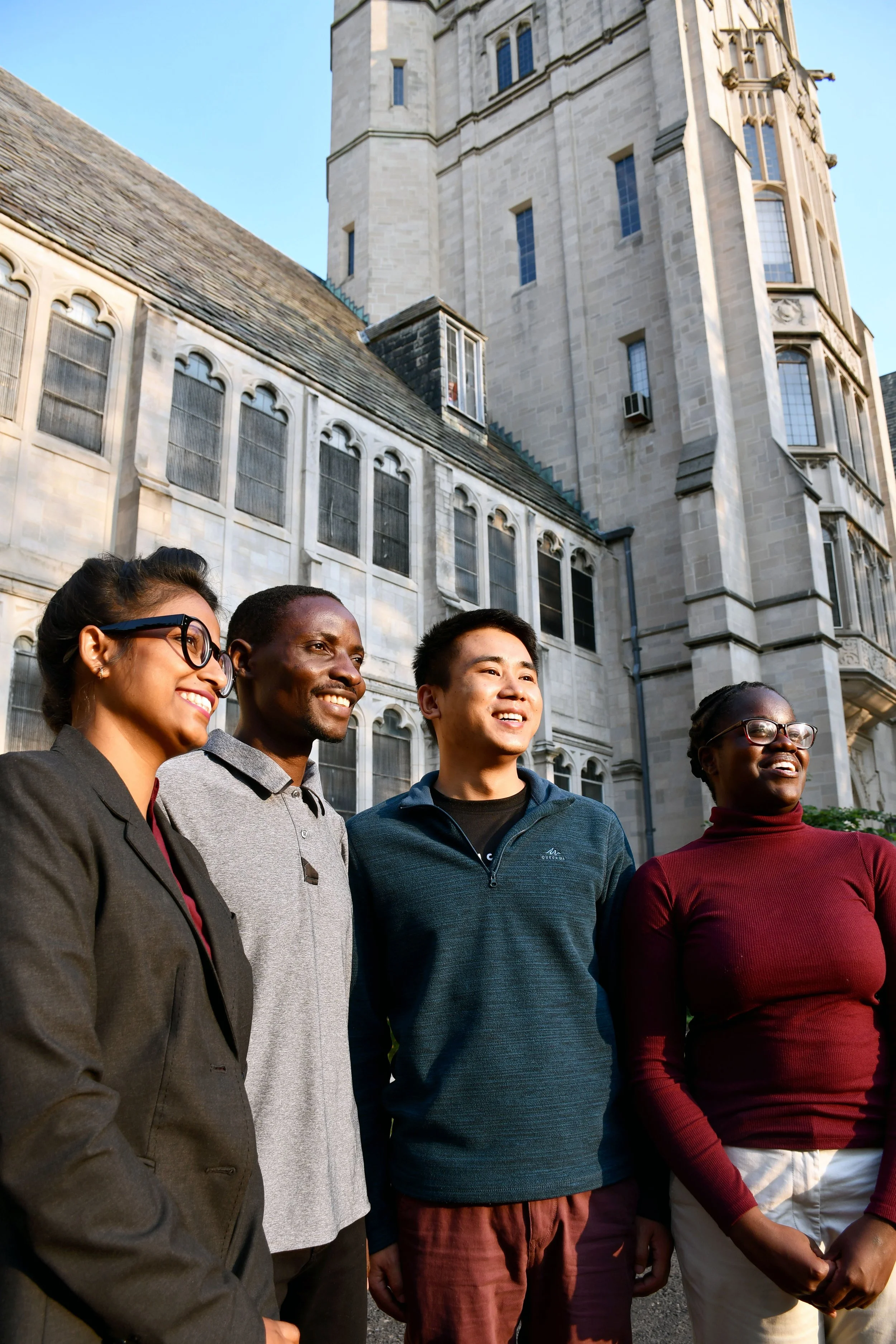 A diverse group of five people standing outdoors in front of a historic stone building with large windows and a tall tower, smiling and looking in the same direction during daylight.