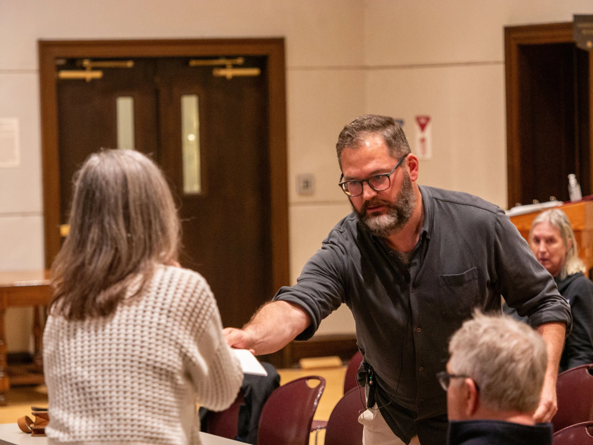 A man with glasses and a beard shaking hands with a woman with gray hair, in a room with wooden doors and seated people.