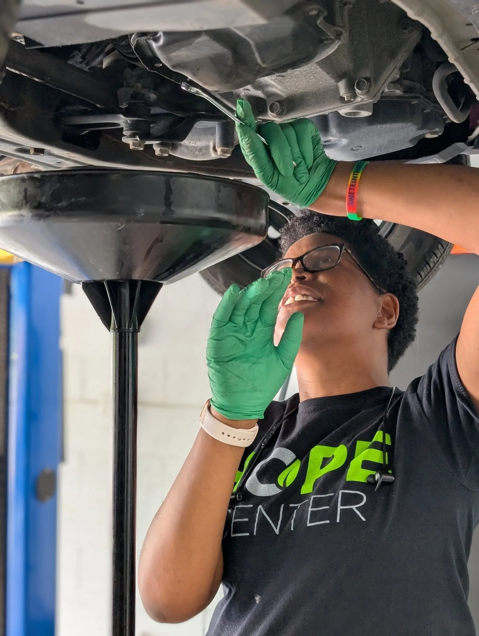 A woman with glasses, wearing a wristband and a gray T-shirt, is working underneath a car, using a tool to work on the car's underbelly.