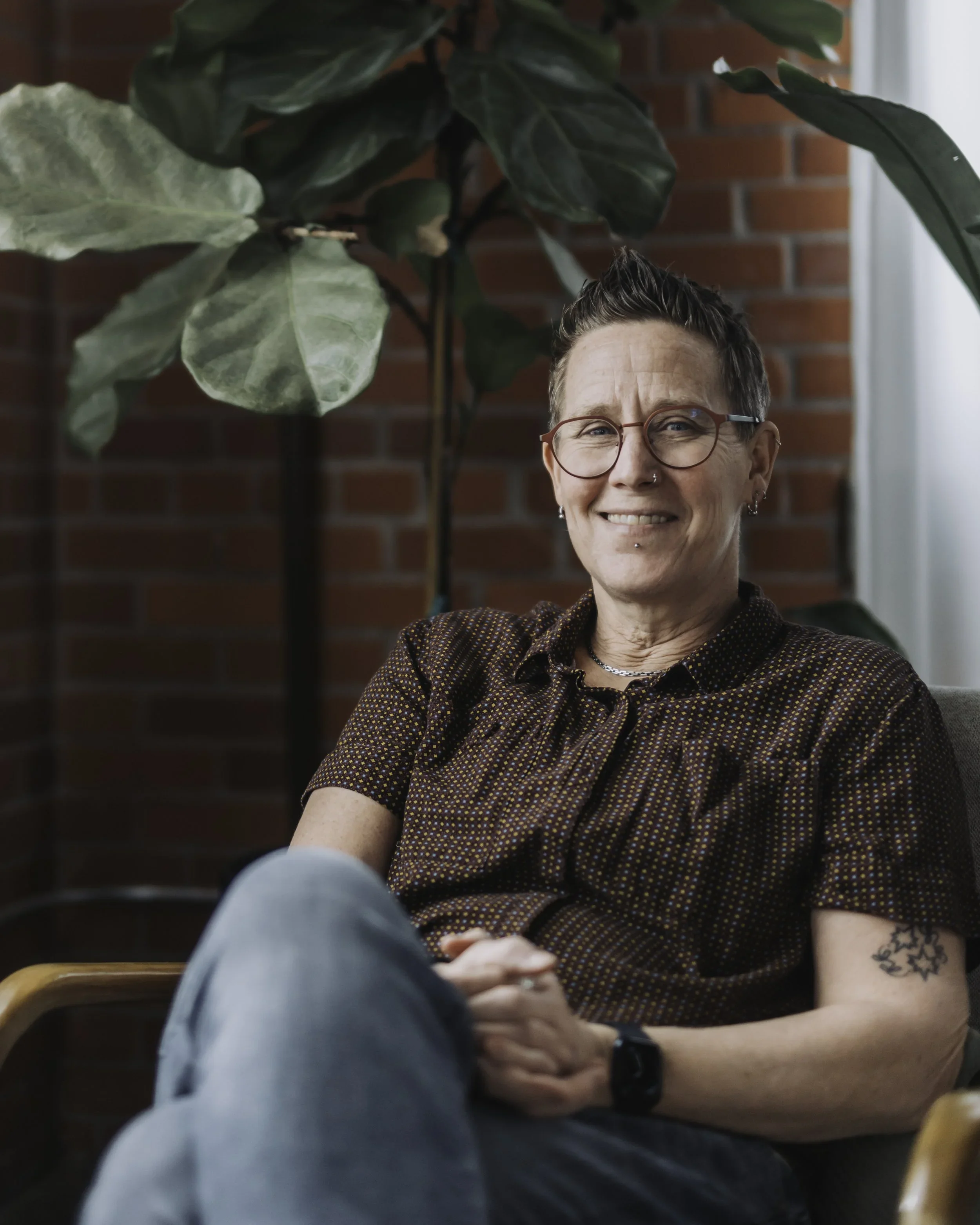 A woman with short hair, glasses, and multiple facial piercings sitting at a table, smiling, with a large leafy plant behind her and a brick wall in the background.