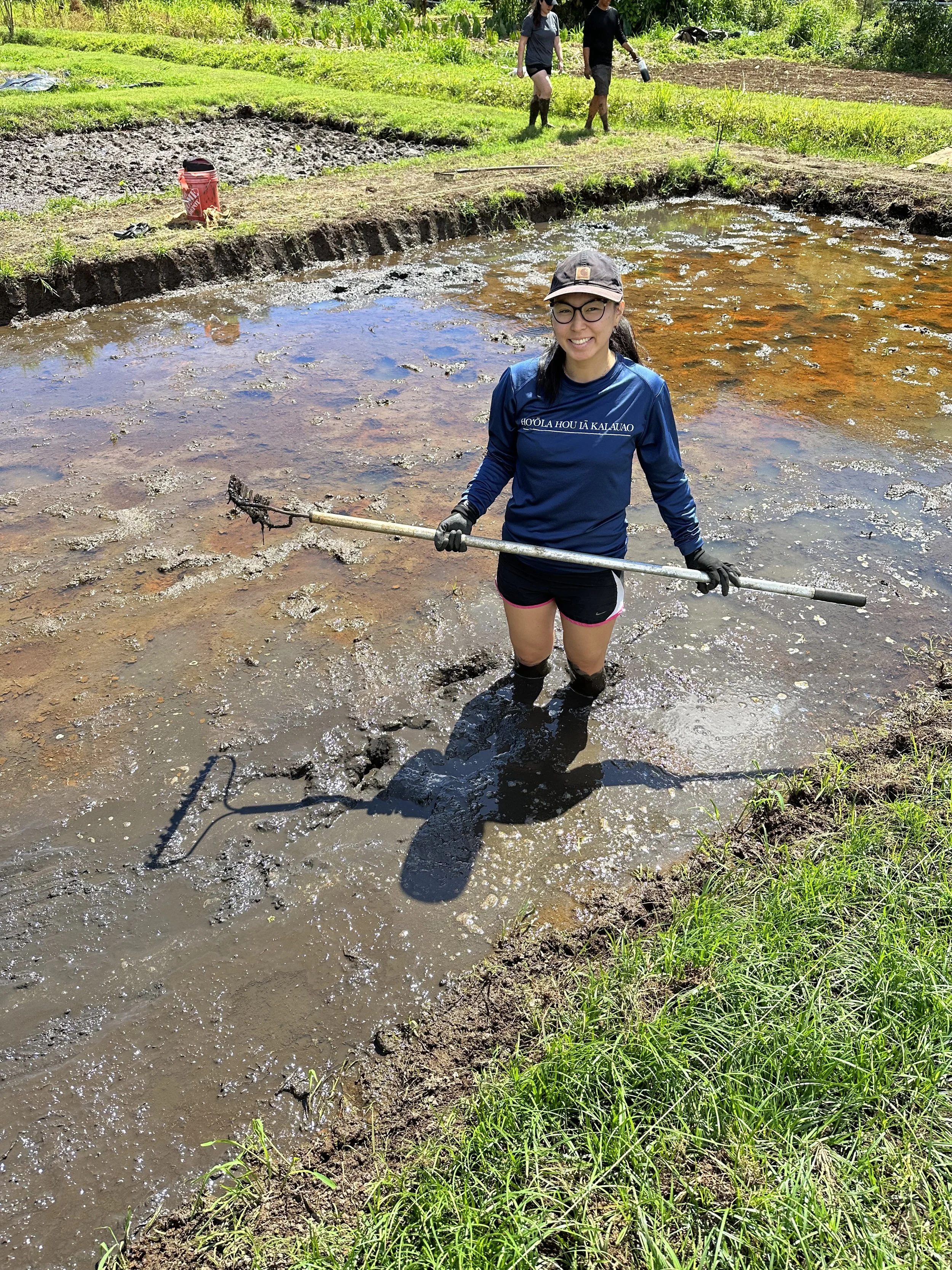 Person smiling and holding a tool while standing in a muddy water-filled agricultural field with two other people working in the background.