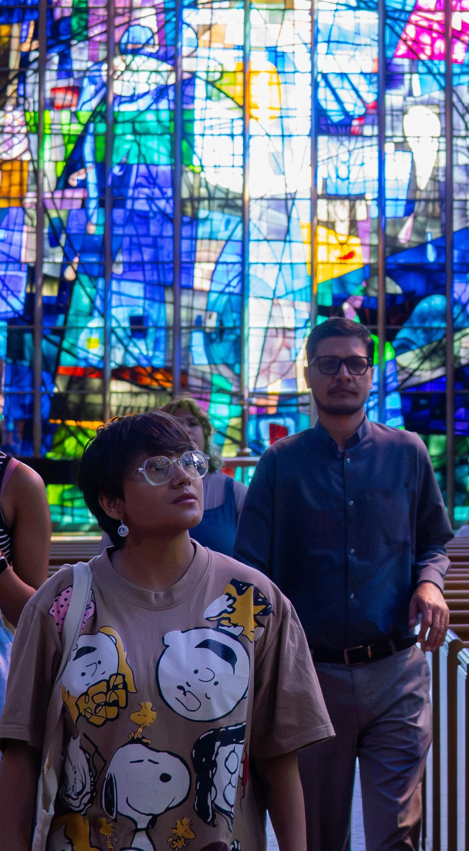 Two people stand in front of a colorful stained glass window in a church or cathedral, with other people partially visible in the background. The woman in the foreground wears glasses, a beige shirt with Peanuts characters, and carries a beige bag, while the man behind her wears glasses, a dark button-up shirt, and beige pants.