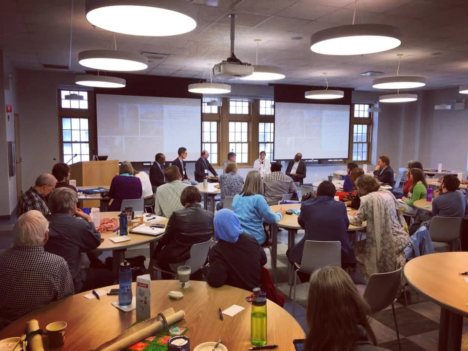 A group of people in a conference room attending a presentation or discussion, seated around round tables with notebooks, drinks, and personal items.