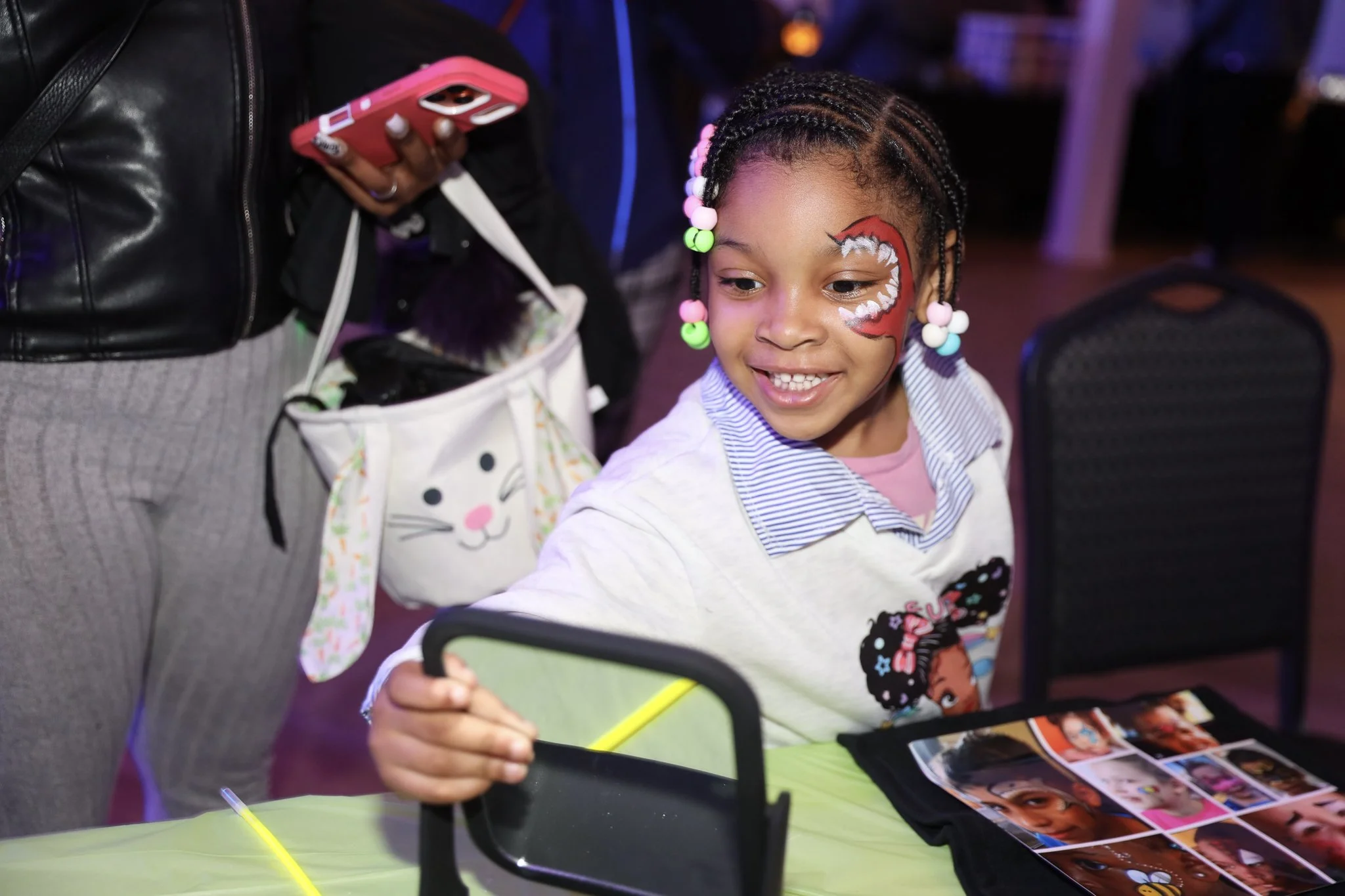 A young girl with braided hair decorated with colorful beads is smiling while looking at photographs on a table, with face paint of a red and white dragon on her cheek.