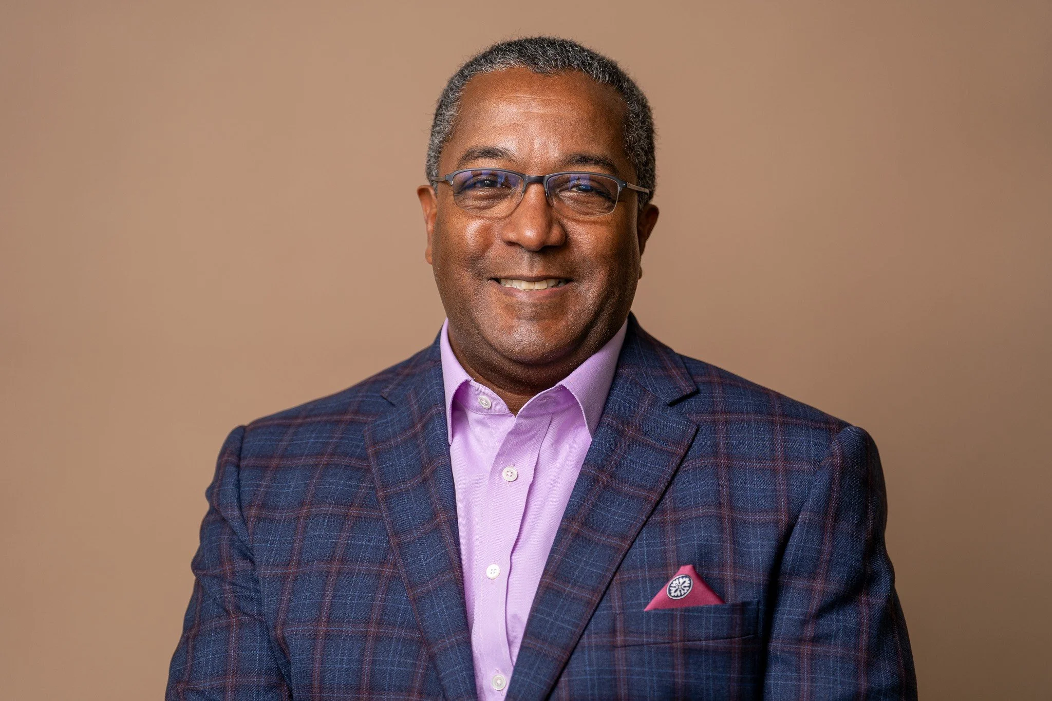 Portrait of an African American man wearing glasses, smiling, dressed in a dark plaid suit with a pink shirt, standing against a beige background.