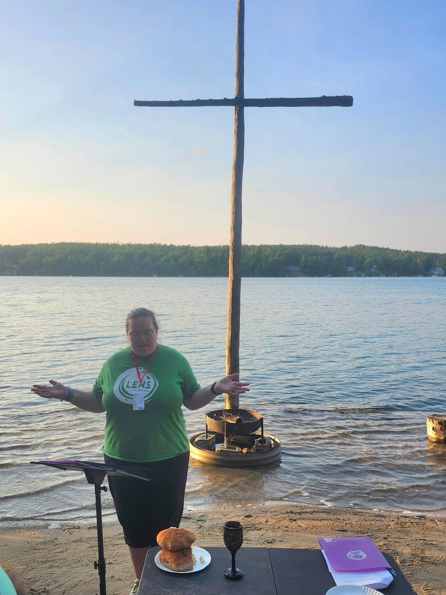 A woman in a green shirt stands in front of a lake at sunset, holding a wooden cross. There is a table with food and drinks, including bread and a purple notebook. The background features trees across the lake and a grill behind the woman.