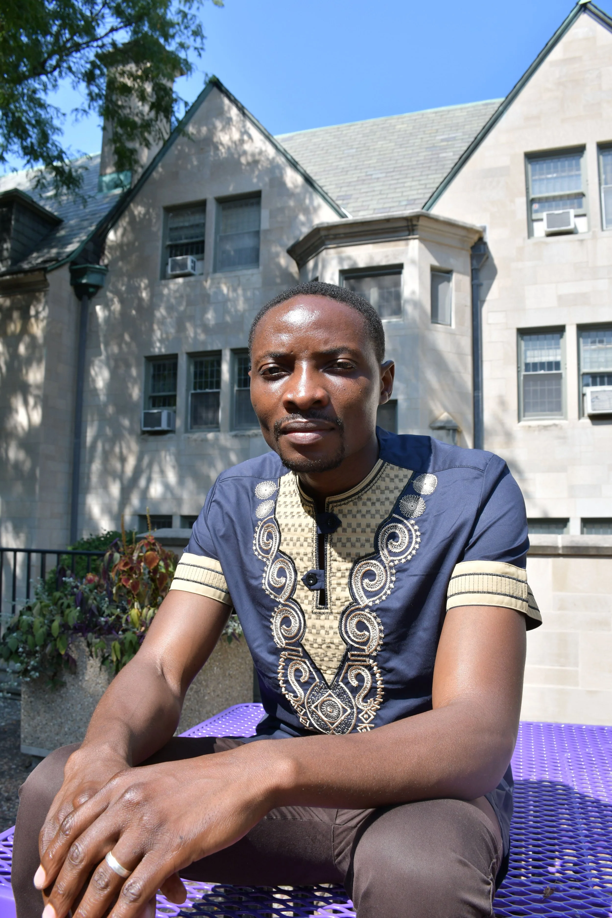 A man wearing a traditional African embroidered shirt sitting outdoors on a purple bench in front of a large stone building with several windows and air conditioning units. There are trees and plants around him, and the sky is clear and blue.