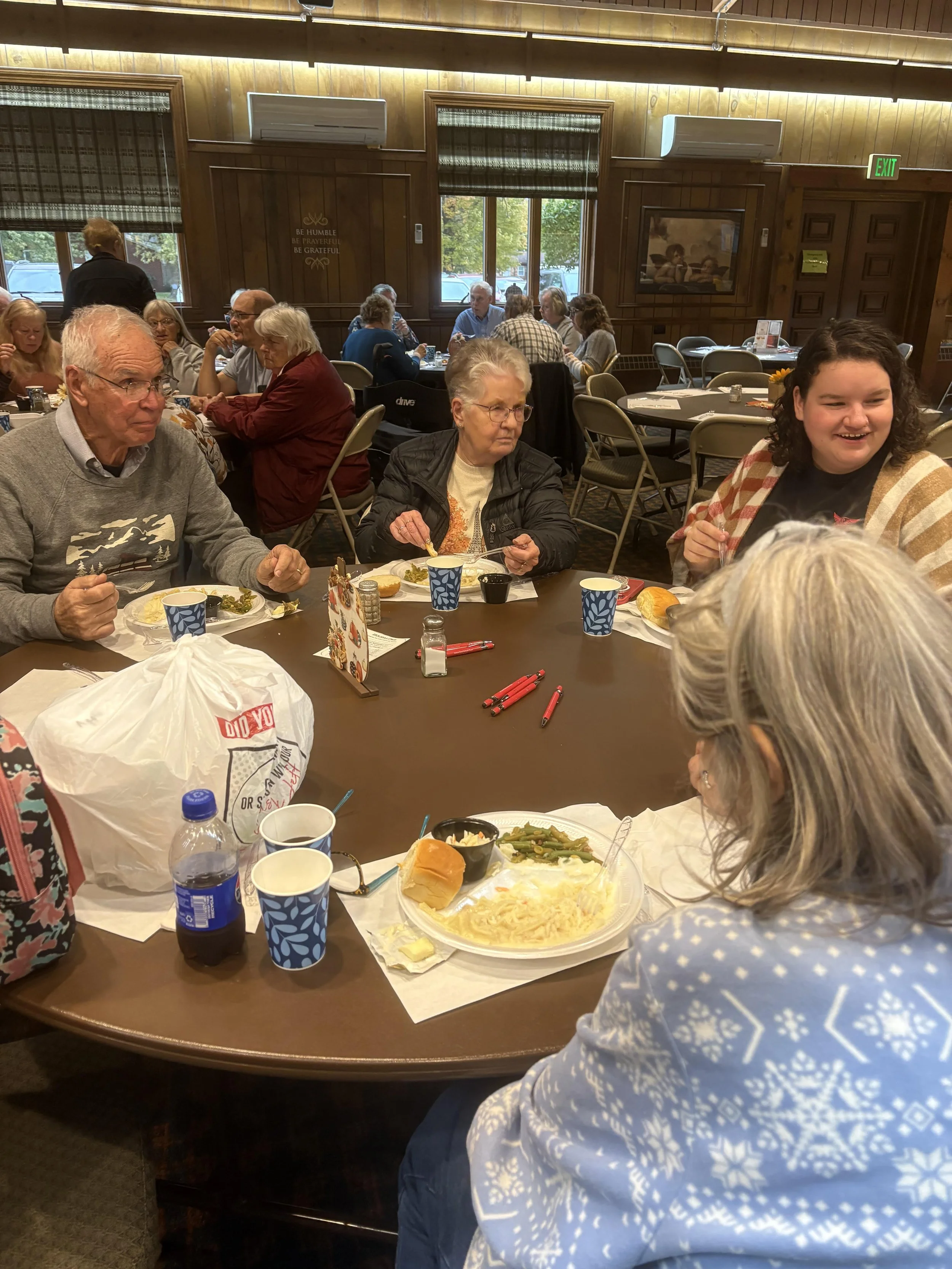 People gathered around a table in a restaurant, enjoying a meal. The table has plates of food, cups, and utensils. The restaurant has wood-paneled walls, windows with blinds, and a wall-mounted TV. A man is in the foreground wearing a gray sweater, and a young woman with curly hair and a striped jacket is smiling on the right side of the image.
