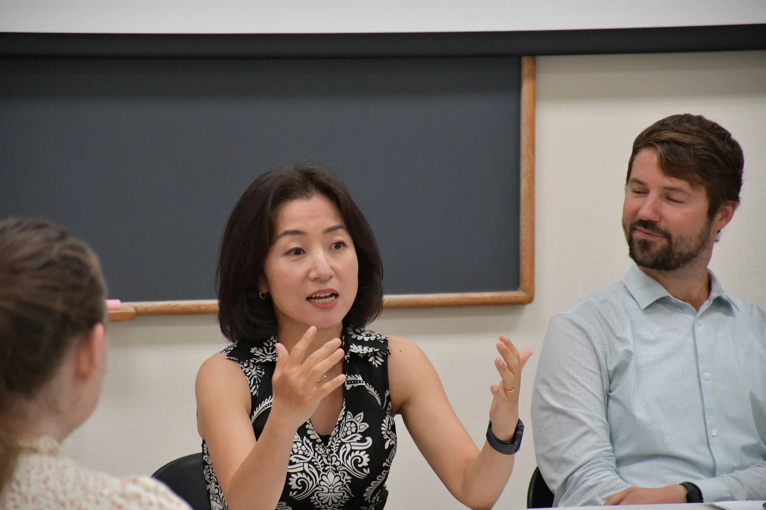 A woman in a black and white patterned dress speaks and gestures with her hands during a discussion, seated at a table with several other people in a classroom or meeting room. A man in a light blue shirt is listening attentively.