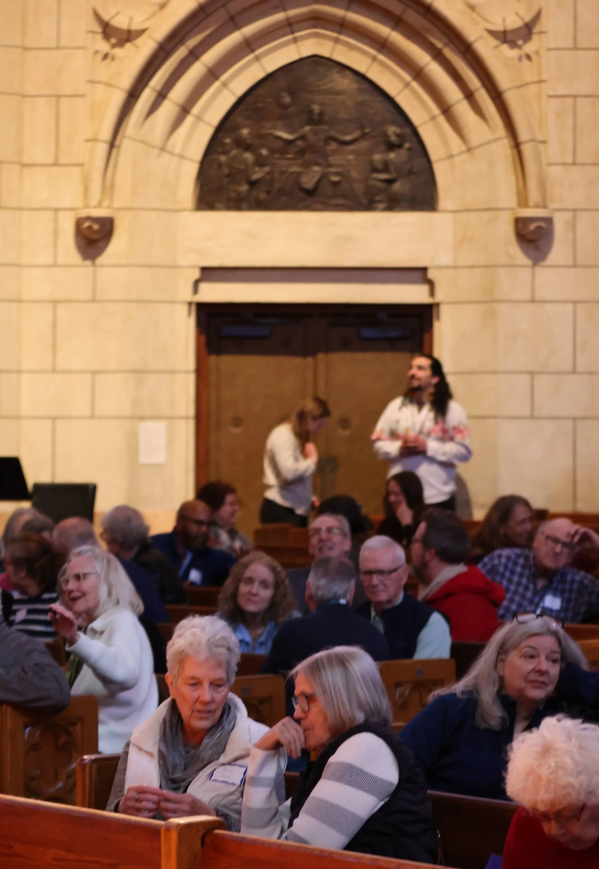 A group of people sitting in a large hall during an event, with two women standing and talking near the wall at the back.