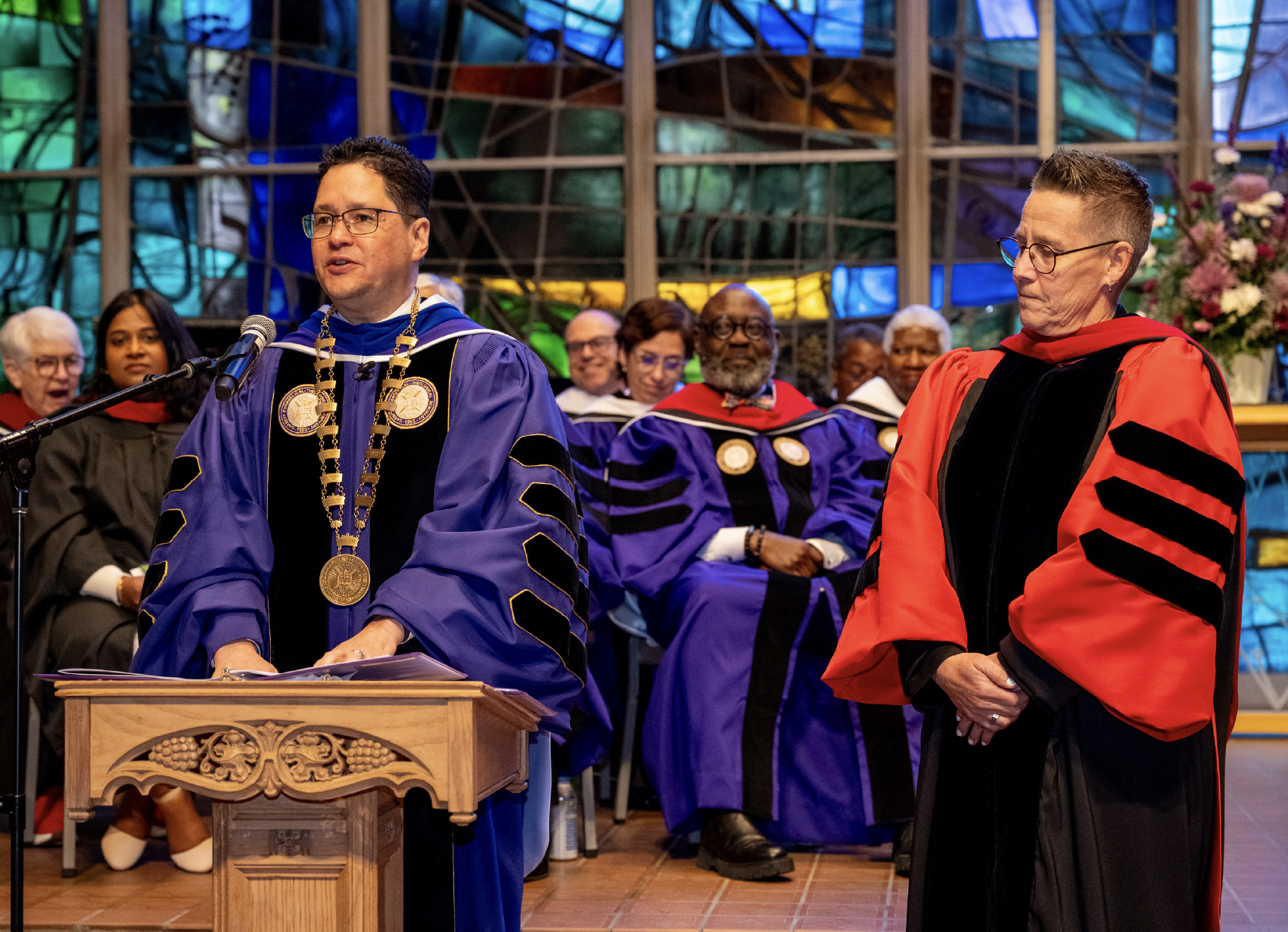 A man in academic regalia speaking at a podium during a graduation ceremony, with several other individuals in academic robes seated behind him inside a building with a colorful stained glass window.