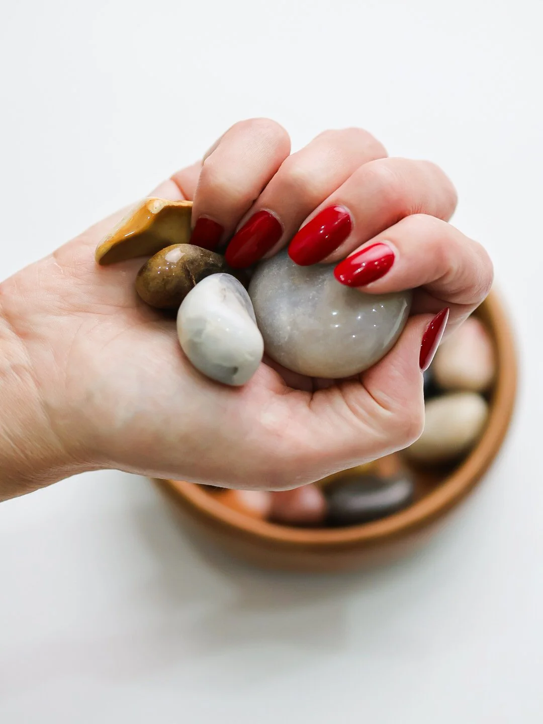 A hand with red painted nails holding several smooth stones, with a bowl of more stones in the background.