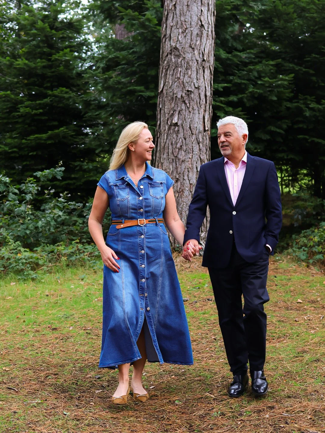 A couple walking hand in hand outdoors in a wooded park, smiling at each other.