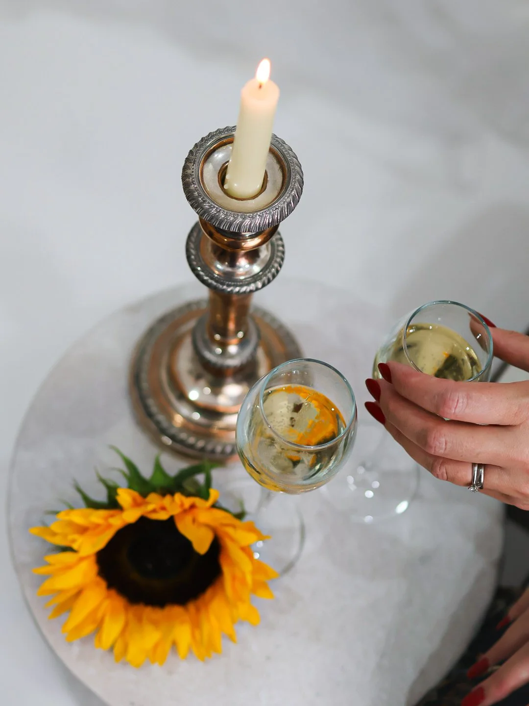 A candle on a silver candle holder with two glasses of white wine, a sunflower, and a hand with red nail polish and a wedding ring on a white table.