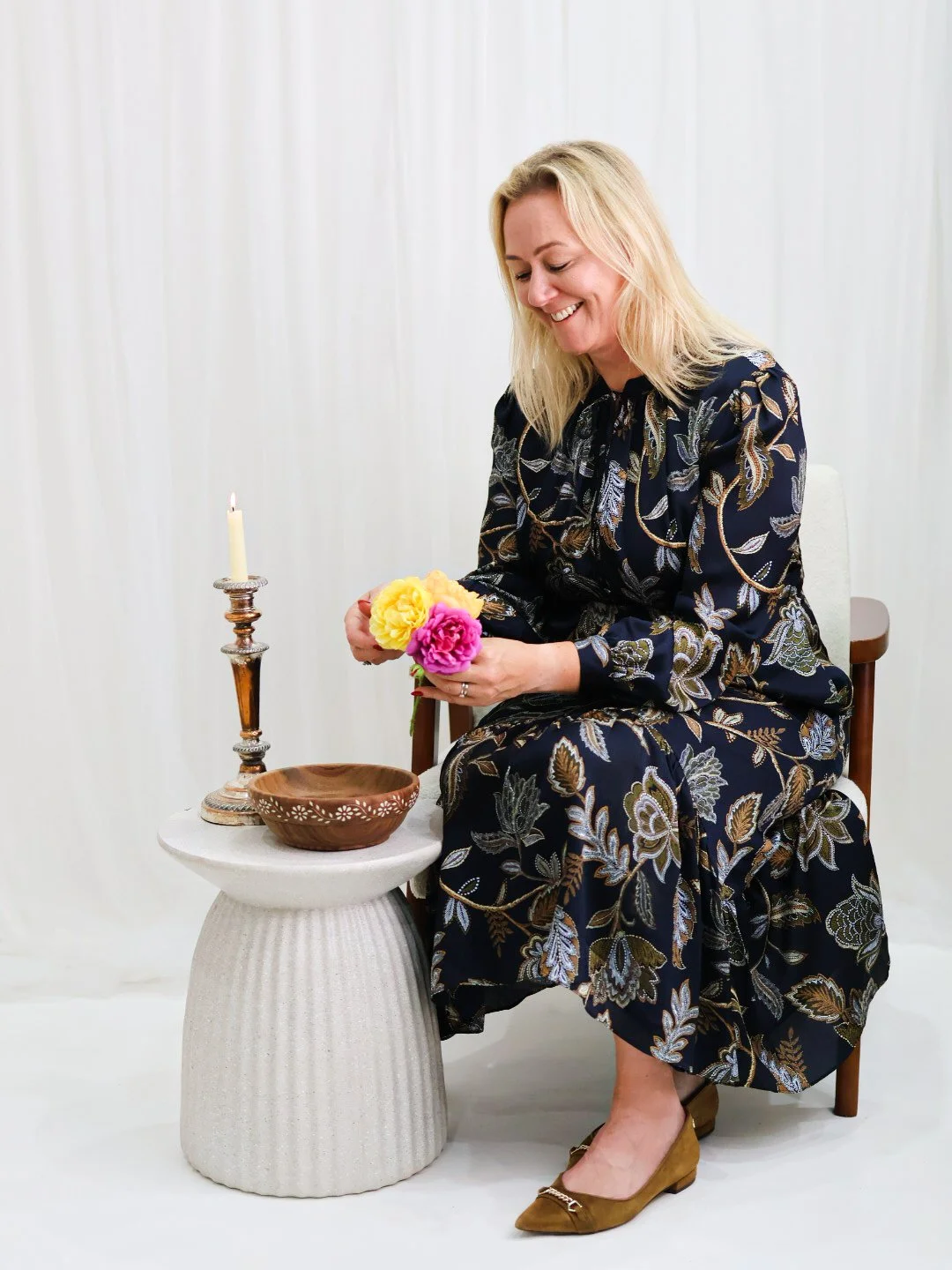 A woman with blonde hair, wearing a dark patterned dress, sits on a chair holding yellow and pink flowers. She is smiling and looking down. There is a candle in a holder, a brown bowl, and a small white table beside her.