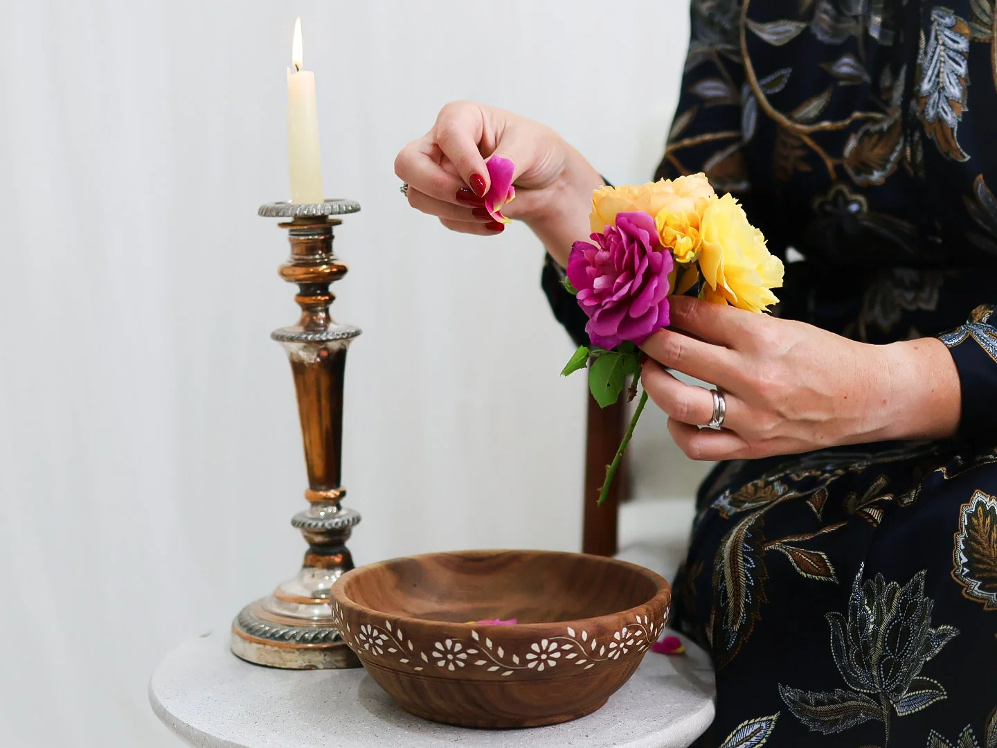 Person arranging pink and yellow flowers in a small bouquet beside a lit candle on a silver candlestick, with a wooden bowl on a white table.