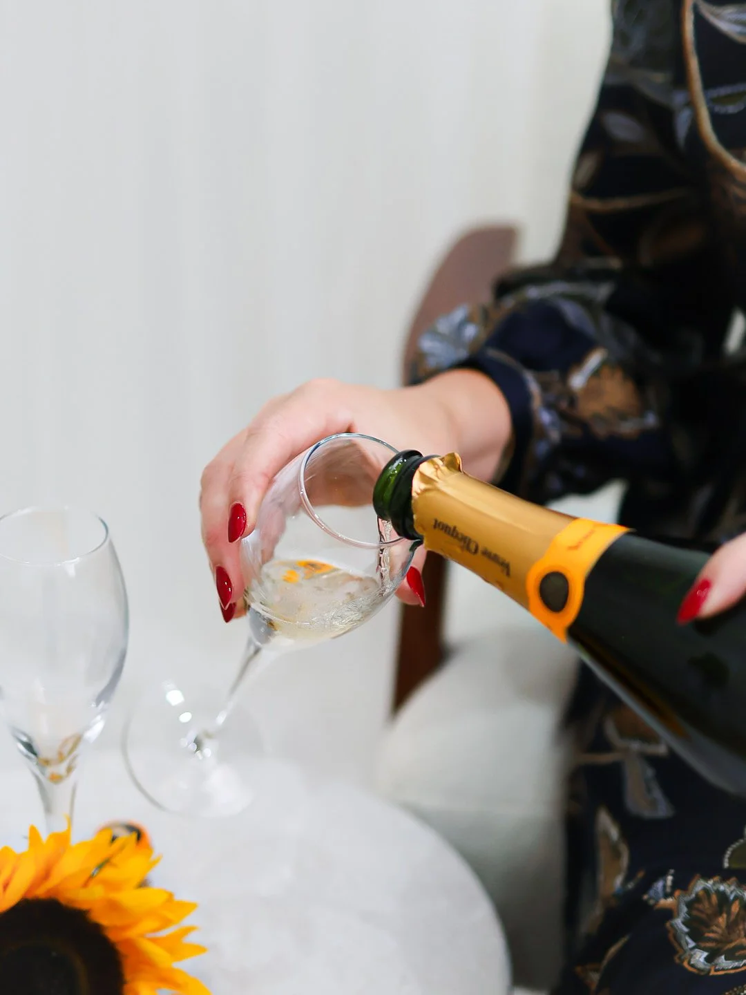 Person wearing patterned clothing pours champagne into a glass, with other empty glasses on the table and a sunflower in the foreground.