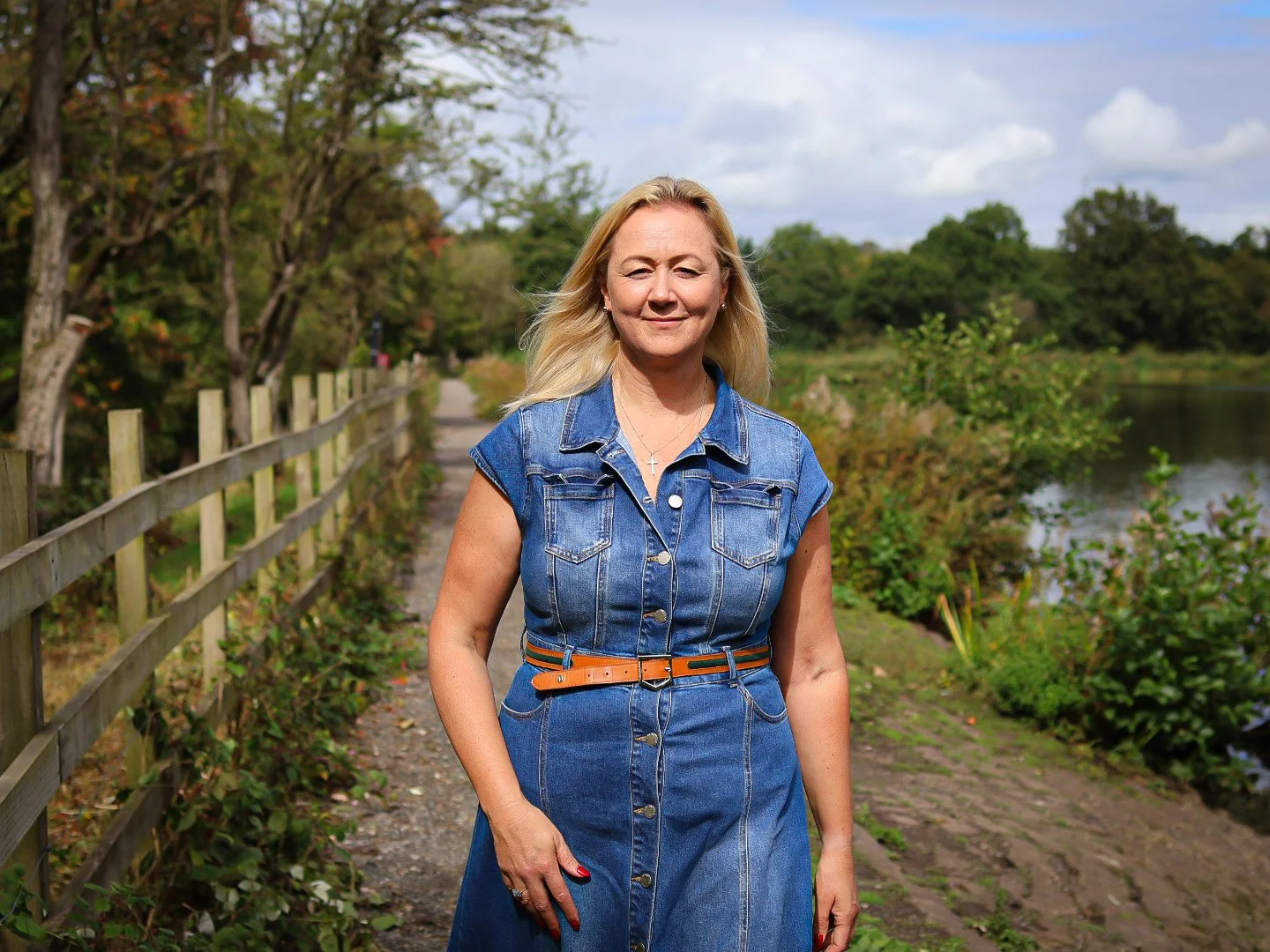 A woman with blonde hair standing on a nature trail beside a body of water, wearing a denim dress with a brown belt and a cross necklace, with trees and blue sky in the background.