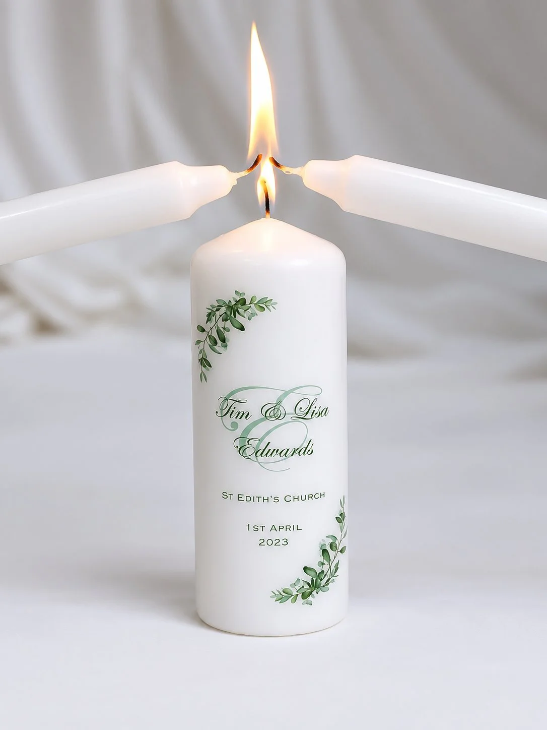 A white memorial candle with green floral decorations and personalized text, three candles are lighting the top of the memorial candle, symbolizing a tribute or remembrance.