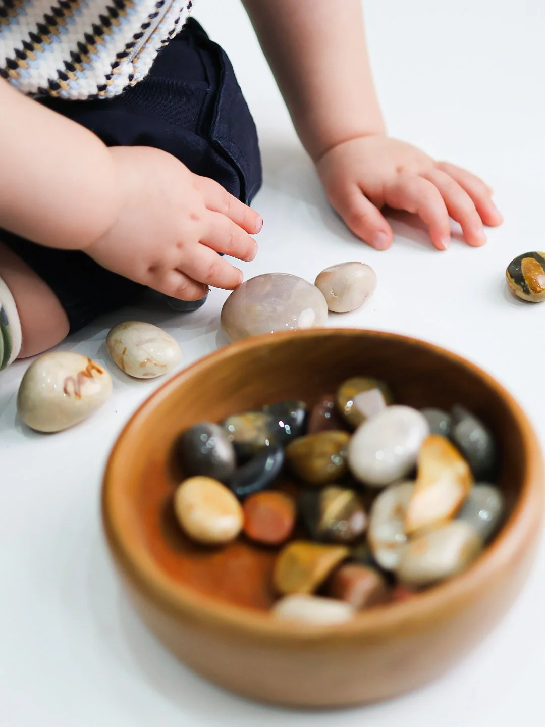 Close-up of a child's hands reaching towards a collection of smooth, polished rocks on a white surface. There is a wooden bowl filled with small, multicolored stones in the foreground.