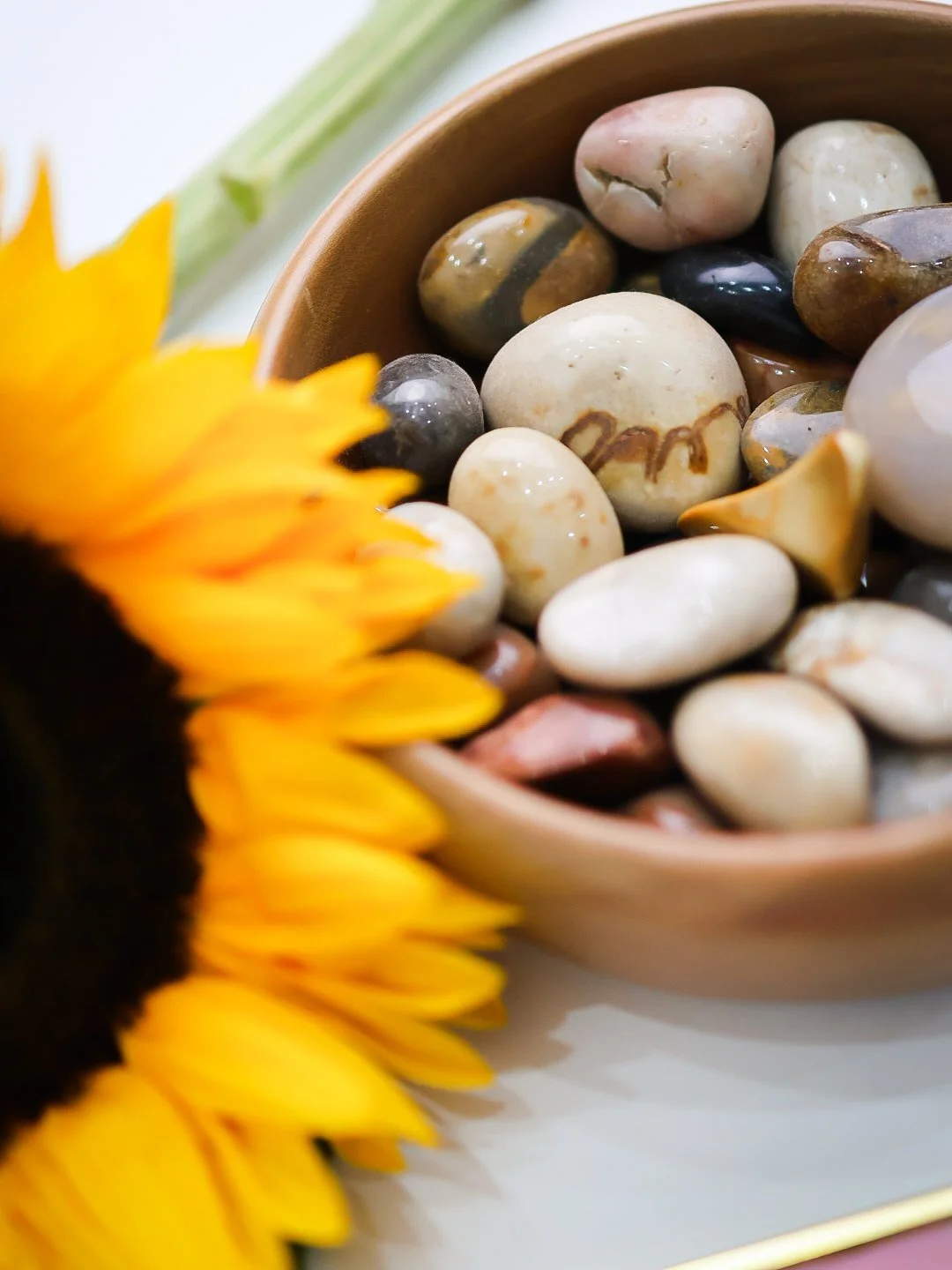 Close-up of a wooden bowl filled with multicolored polished stones and a blurred yellow sunflower in the foreground.