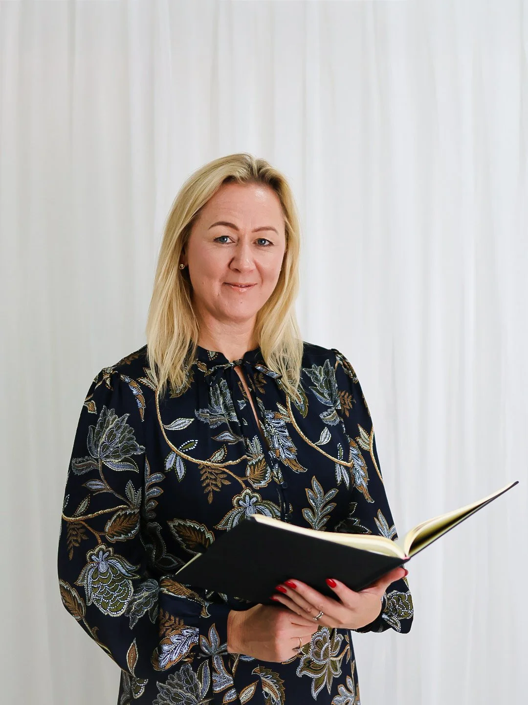 A blonde woman in a black floral dress holding an open book, standing in front of white curtains.