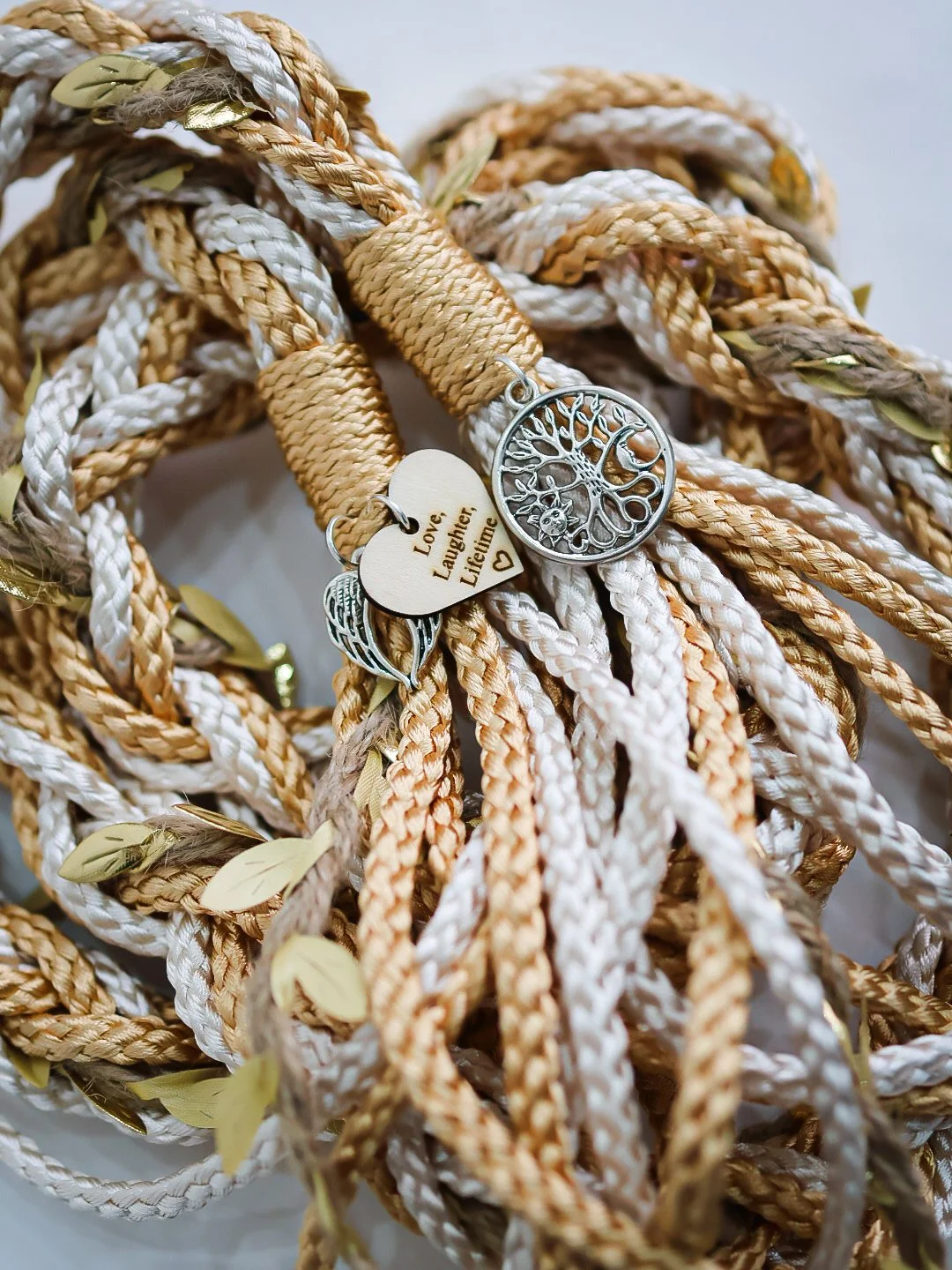 A pile of tangled beige, white, and light brown braided cords decorated with small gold-colored leaves and three small charms, including a heart engraved with "Love, Laughter, Lifetime," a teardrop-shaped charm, and a circular charm with a tree of life design.