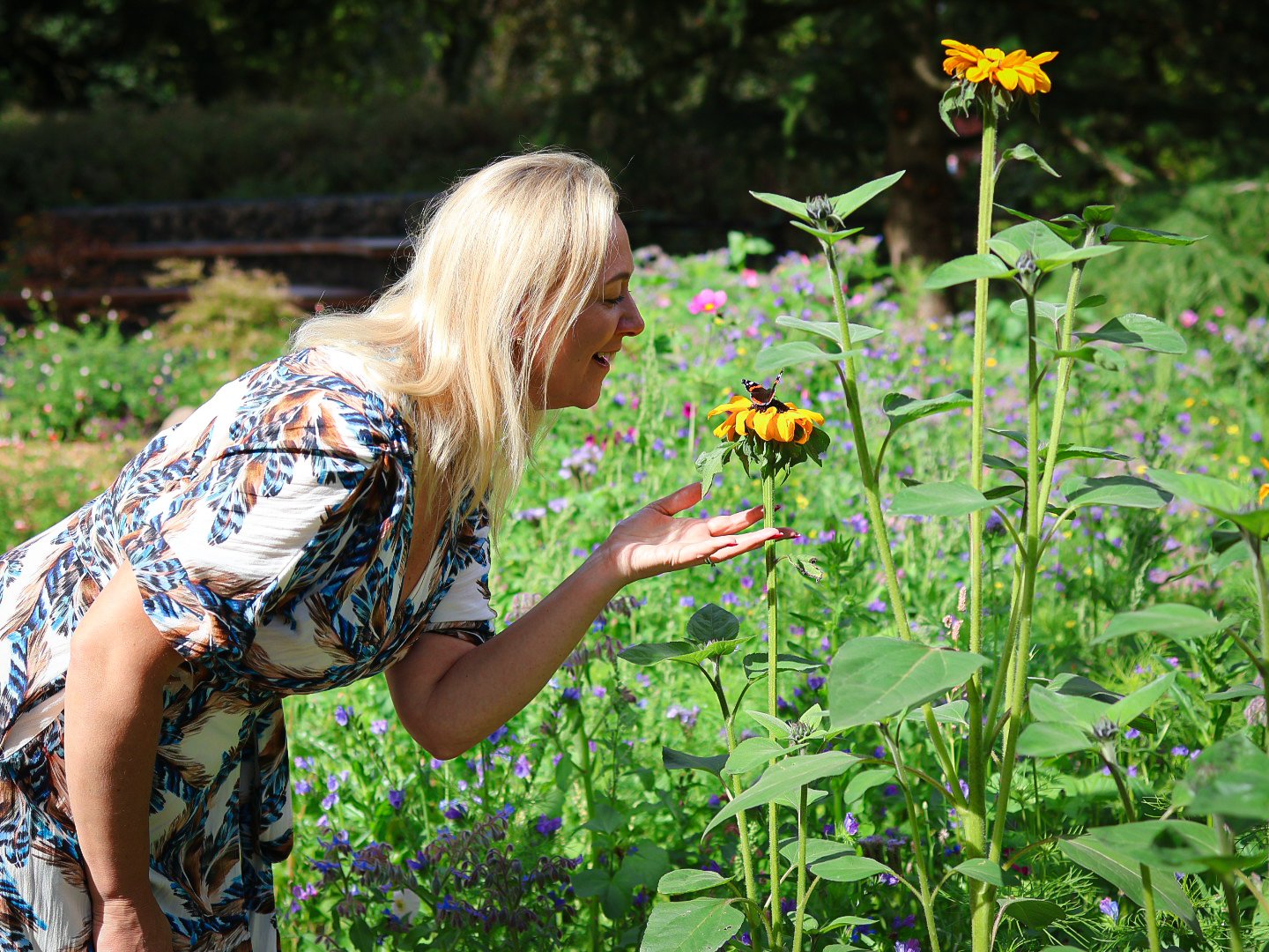 A woman with long blonde hair in a patterned dress is outdoors in a garden with tall sunflower plants. She is leaning forward with her hand extended, looking at a butterfly perched on a sunflower, surrounded by colorful flowers and greenery.